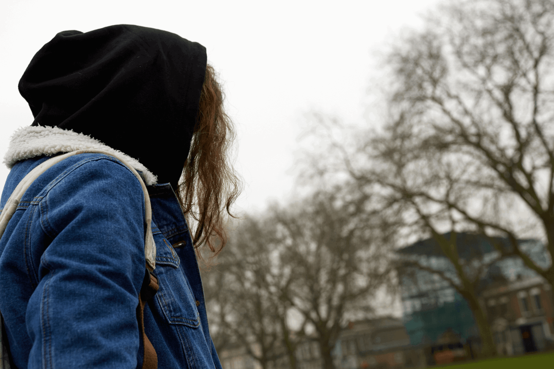 A girl wearing a backpack, denim jacket and hoodie stands in a park with her face turned away.