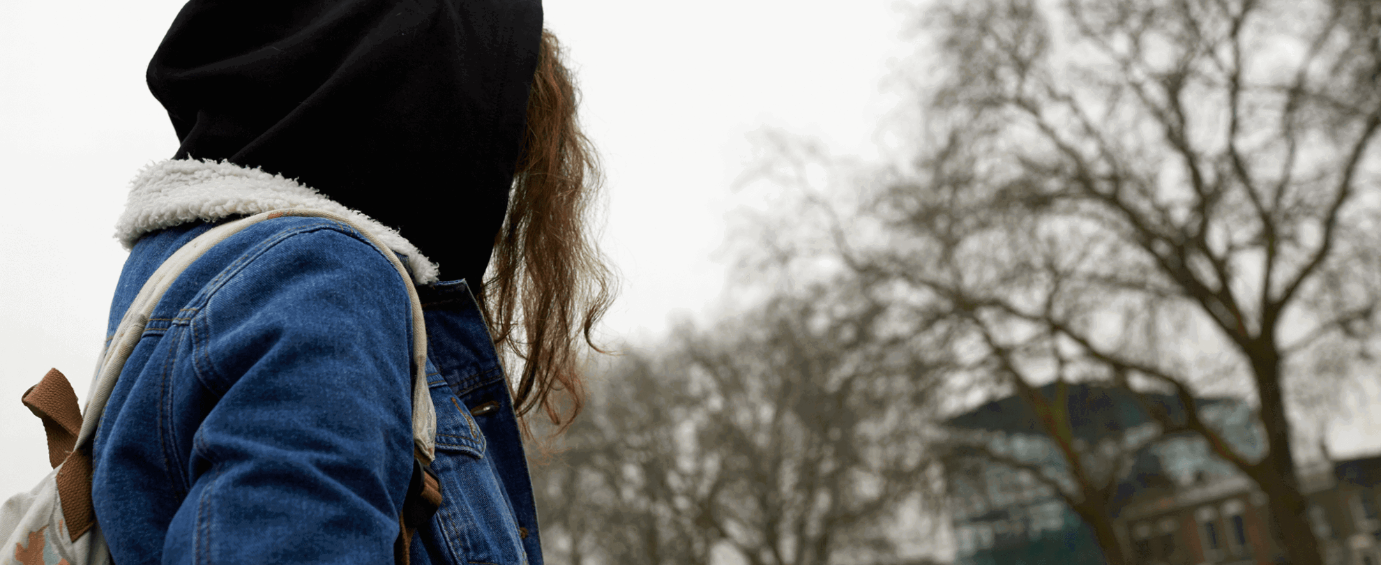 A girl wearing a backpack, denim jacket and hoodie stands in a park with her face turned away.