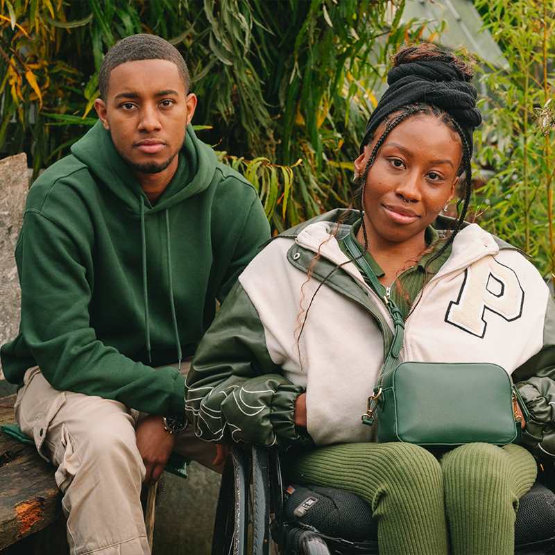 A young Black woman in a wheelchair and a young Black man on a bench, both staring at the camera looking serious.