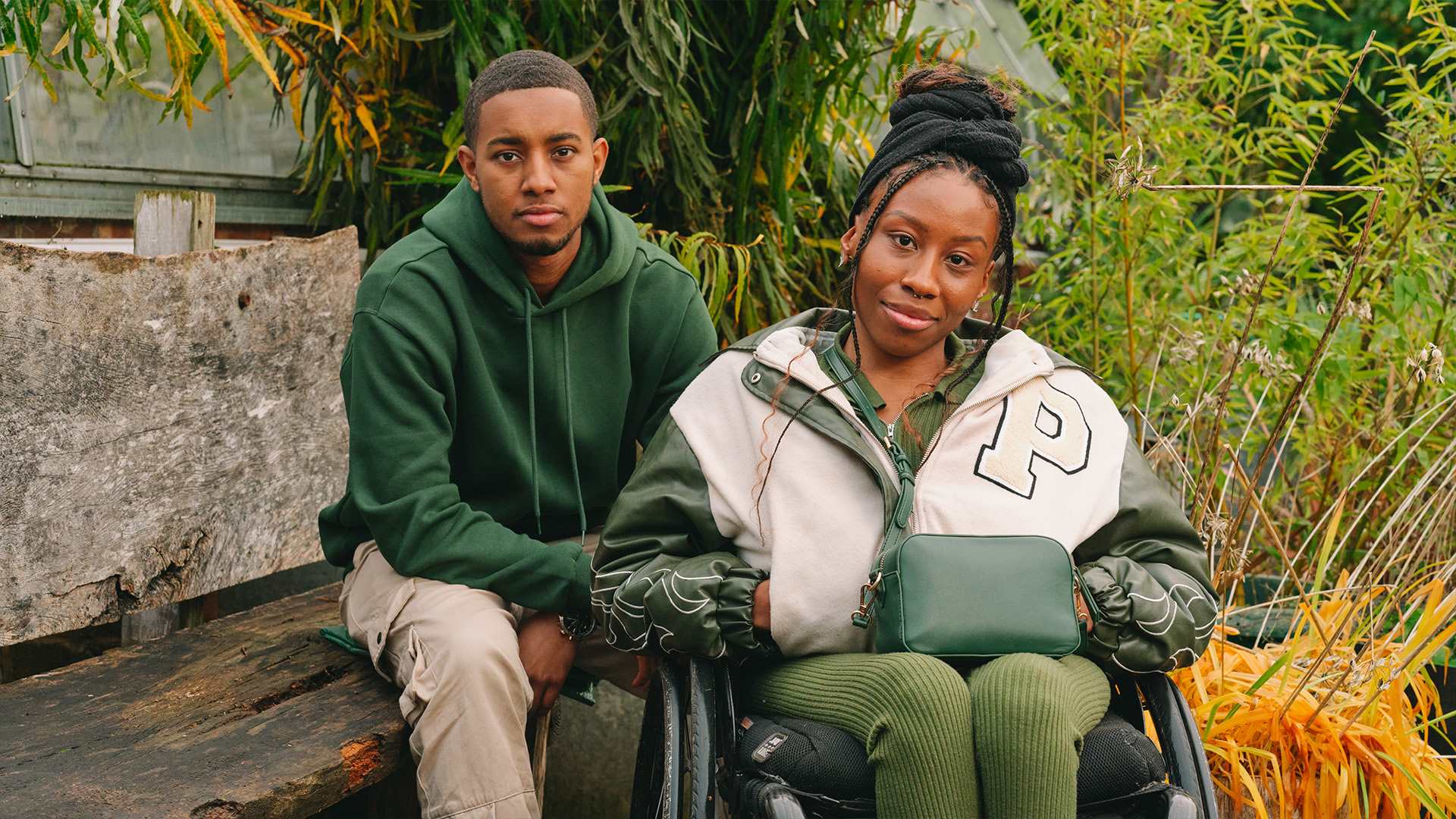 A young Black woman in a wheelchair and a young Black man on a bench, both staring at the camera looking serious.