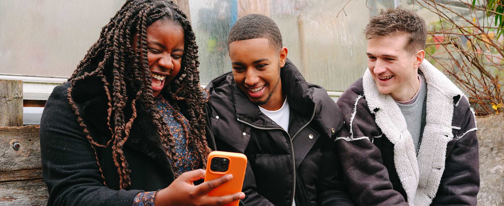 A young Black woman, young Black man and young white man, all sitting on a bench outside, looking at something on a phone and laughing.