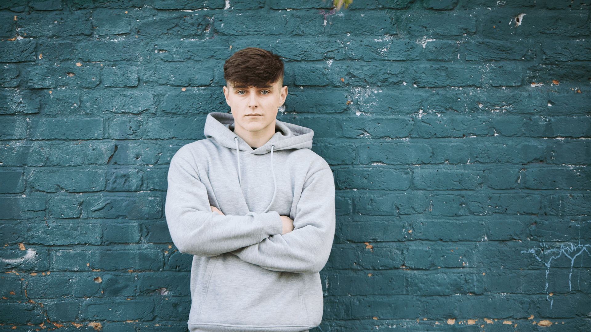 a-boy-wearing-grey-hoodie-with-his-arms-crossed-looking-in-front-of-the-camera-while-leaning-against-a-turquoise-wall