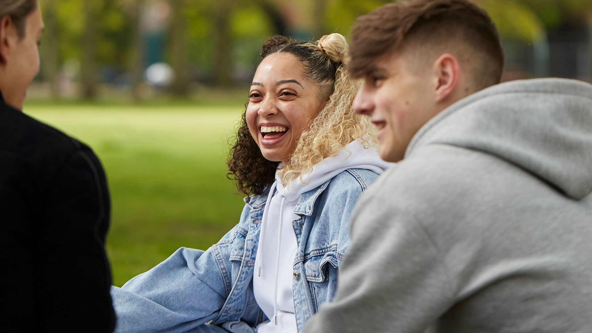 Three young people sit in a park, chatting. The person in the middle has curly hair and wears a denim jacket and is laughing, while looking at the two other young people. The person on the right is wearing a grey hoodie, the person on the left wears a black jacket.