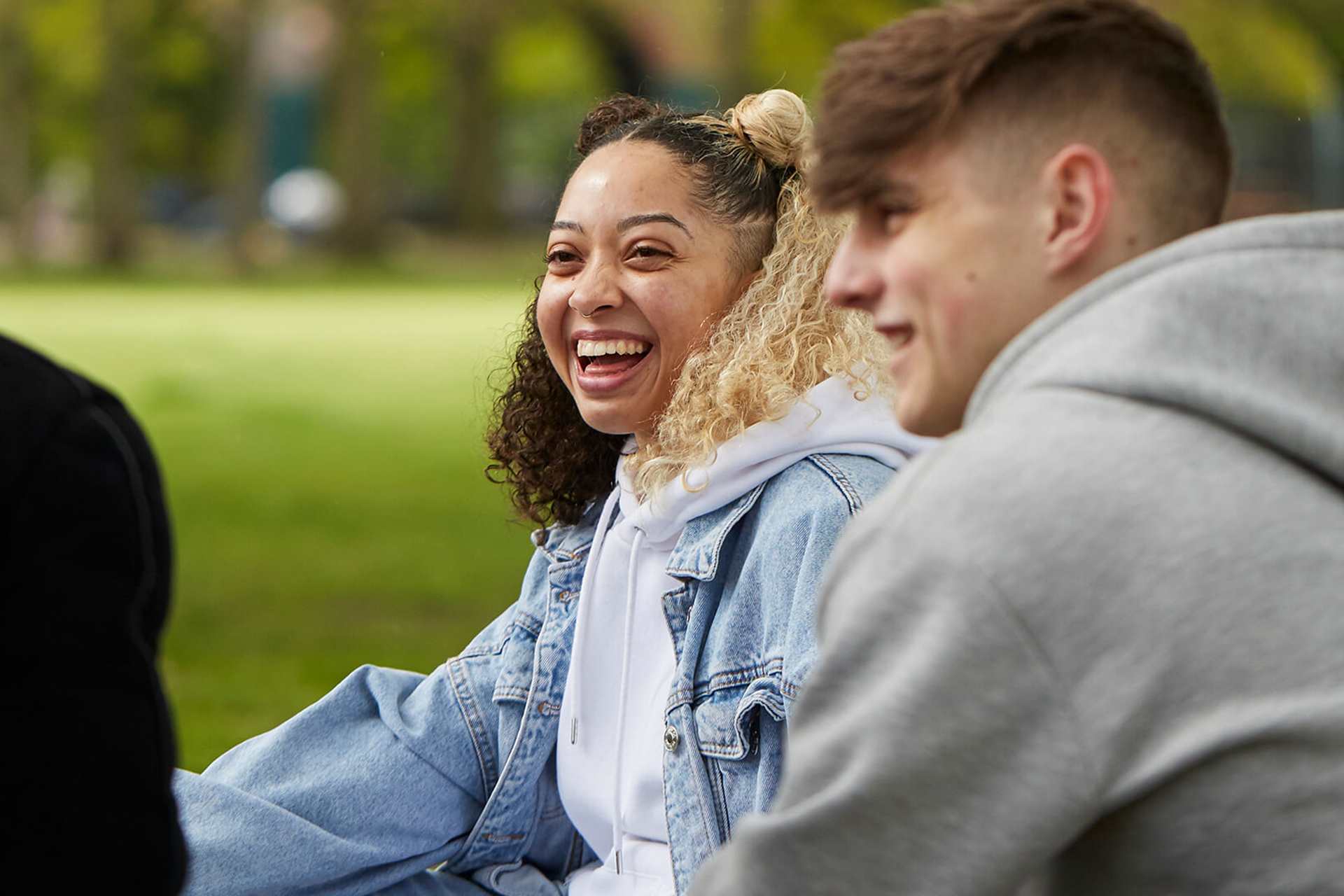 Three young people sit in a park, chatting. The person in the middle has curly hair and wears a denim jacket and is laughing, while looking at the two other young people. The person on the right is wearing a grey hoodie, the person on the left wears a black jacket.