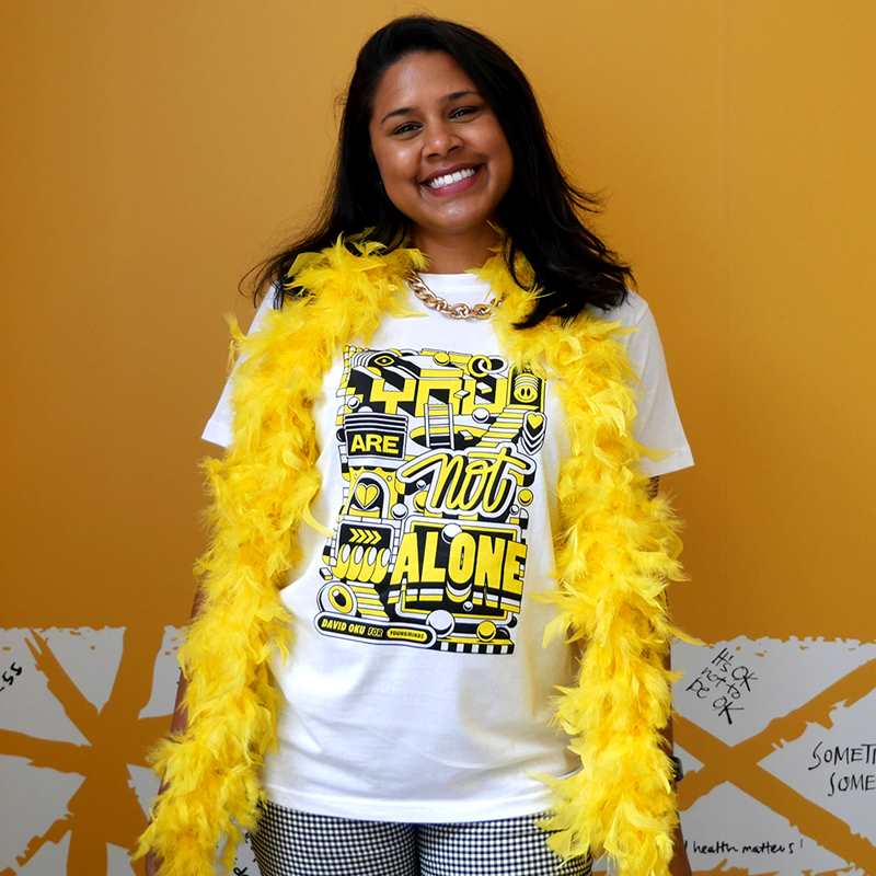 A woman smiling wearing a yellow feather bower, gold chain and our David Oku's bespoke 'You are not alone' graphic #HelloYellow T-shirt.