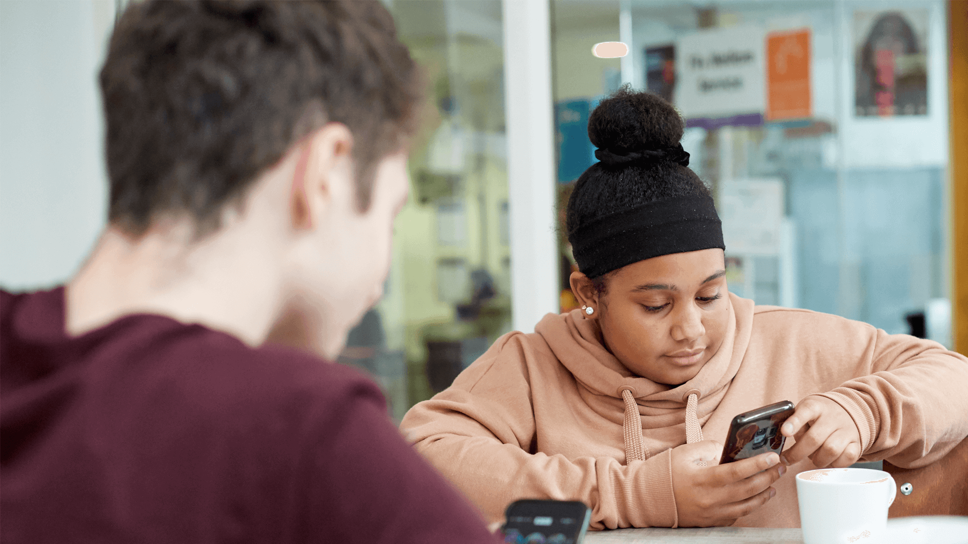 a girl wearing pink hoodie and a boy wearing red shirt in front of her are busy looking at their phones while sitting inside the campus