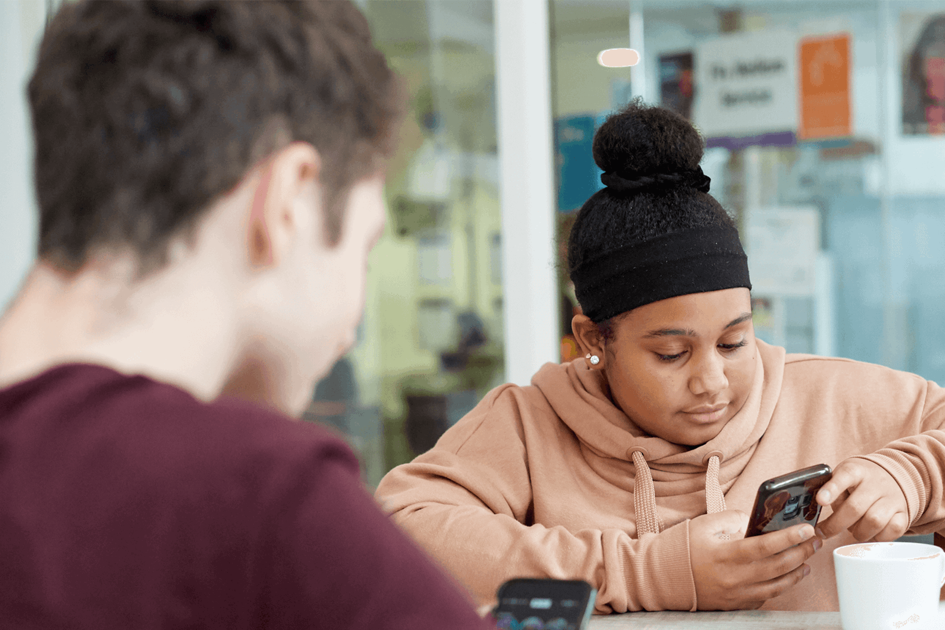 a girl wearing pink hoodie and a boy wearing red shirt in front of her are busy looking at their phones while sitting inside the campus