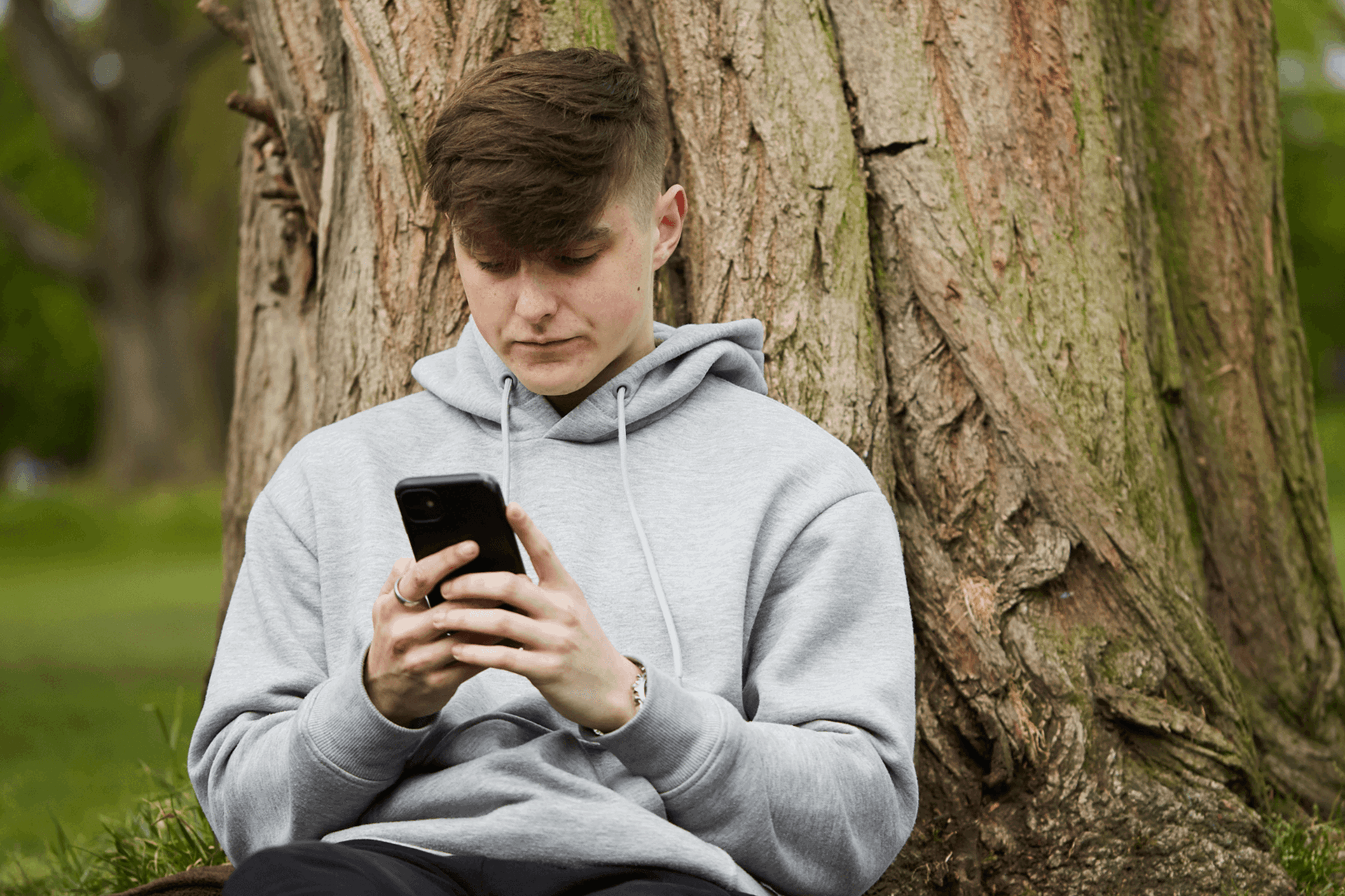 a boy wearing grey hoodie using his mobile phone while sitting on the ground leaning on a tree