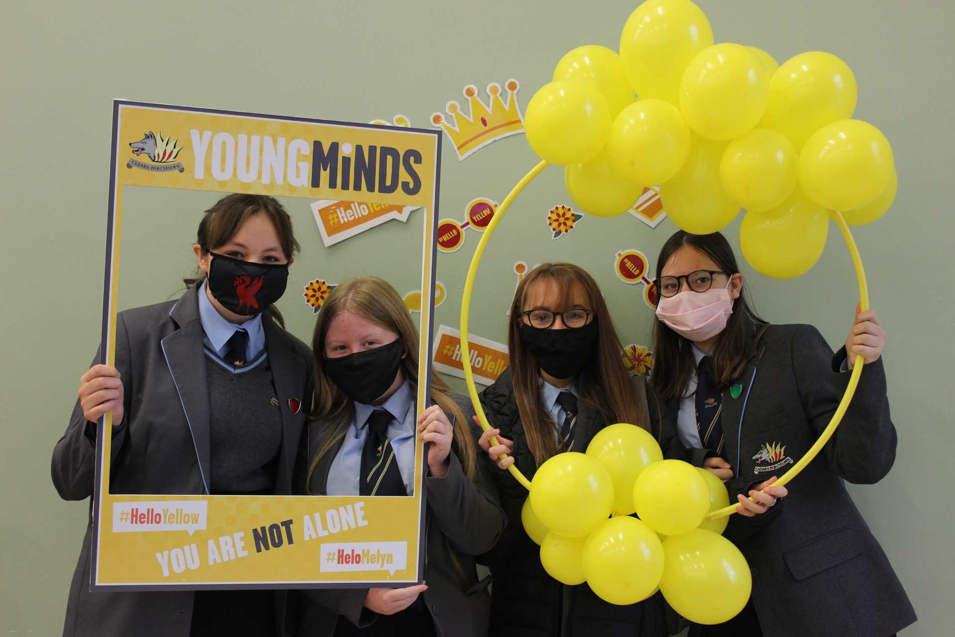 Four students all with masks pose and stand together. Two are in a yellow photo frame and the other two are in a yellow hoop with balloons on.