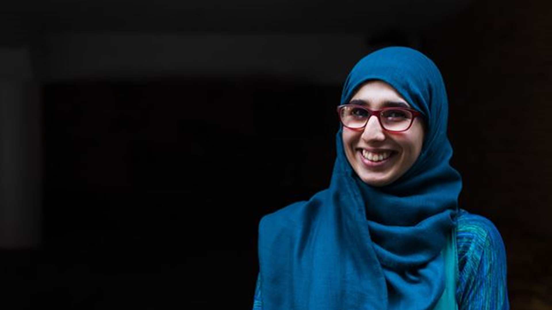 A young woman wearing a blue hijab and glasses smiling.