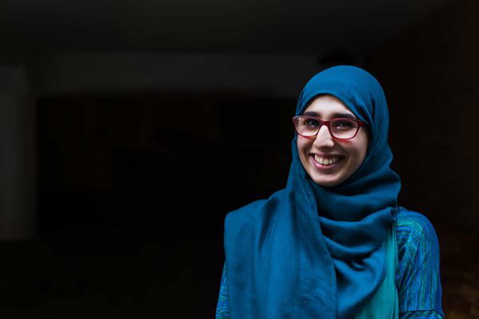 A young woman smiling and wearing a blue hijab and glasses.