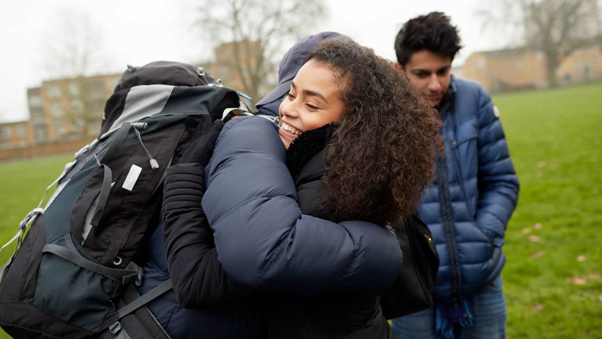 medium-shot-of-a-young-woman-with-curly-hair-smiling-while-hugging-another-person-with-face-unseen-and-wearing-a-cap-blue-jacket-and-a-huge-backpack-with-another-young-man-looking-down-with-school-building-on-background