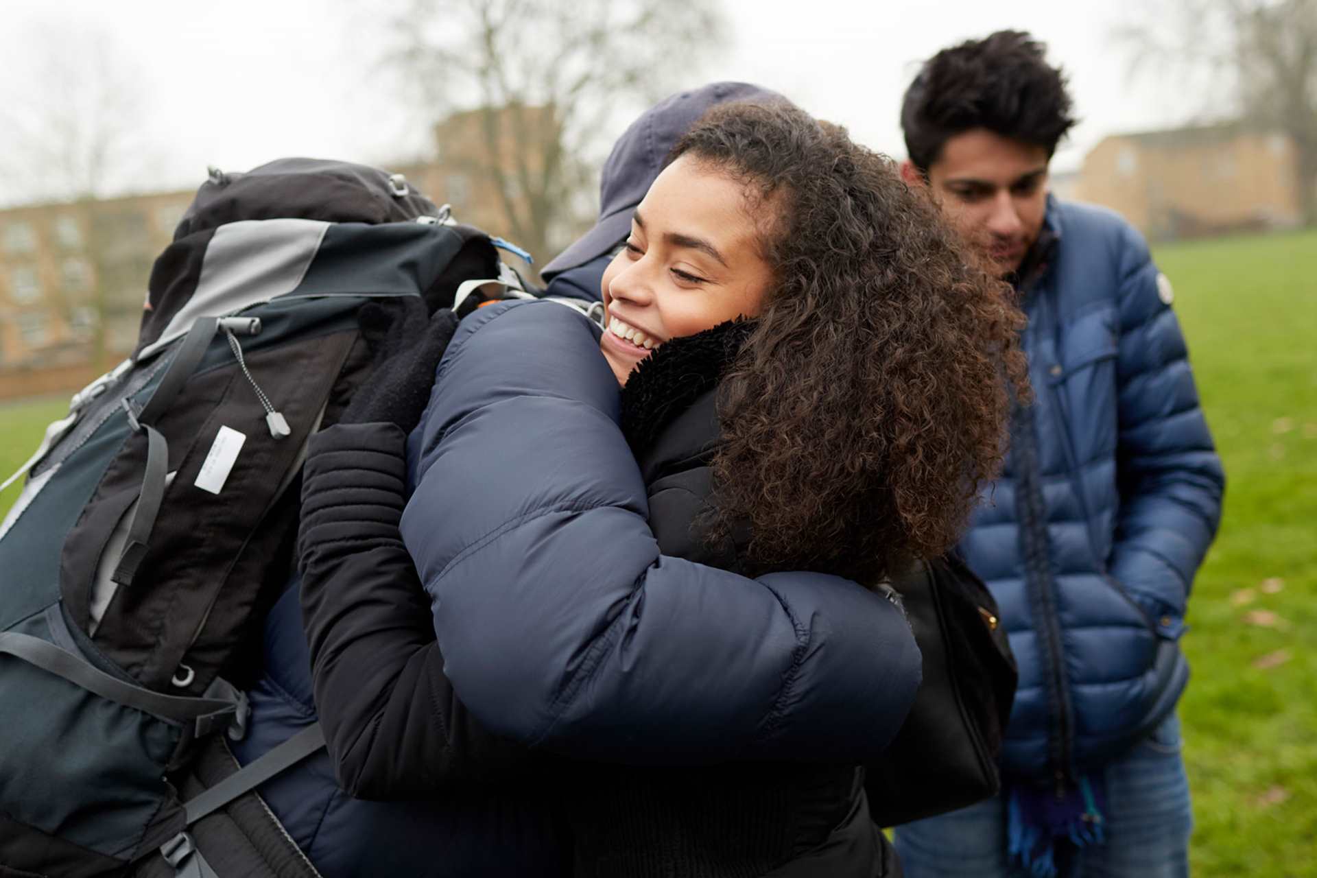 medium-shot-of-a-young-woman-with-curly-hair-smiling-while-hugging-another-person-with-face-unseen-and-wearing-a-cap-blue-jacket-and-a-huge-backpack-with-another-young-man-looking-down-with-school-building-on-background