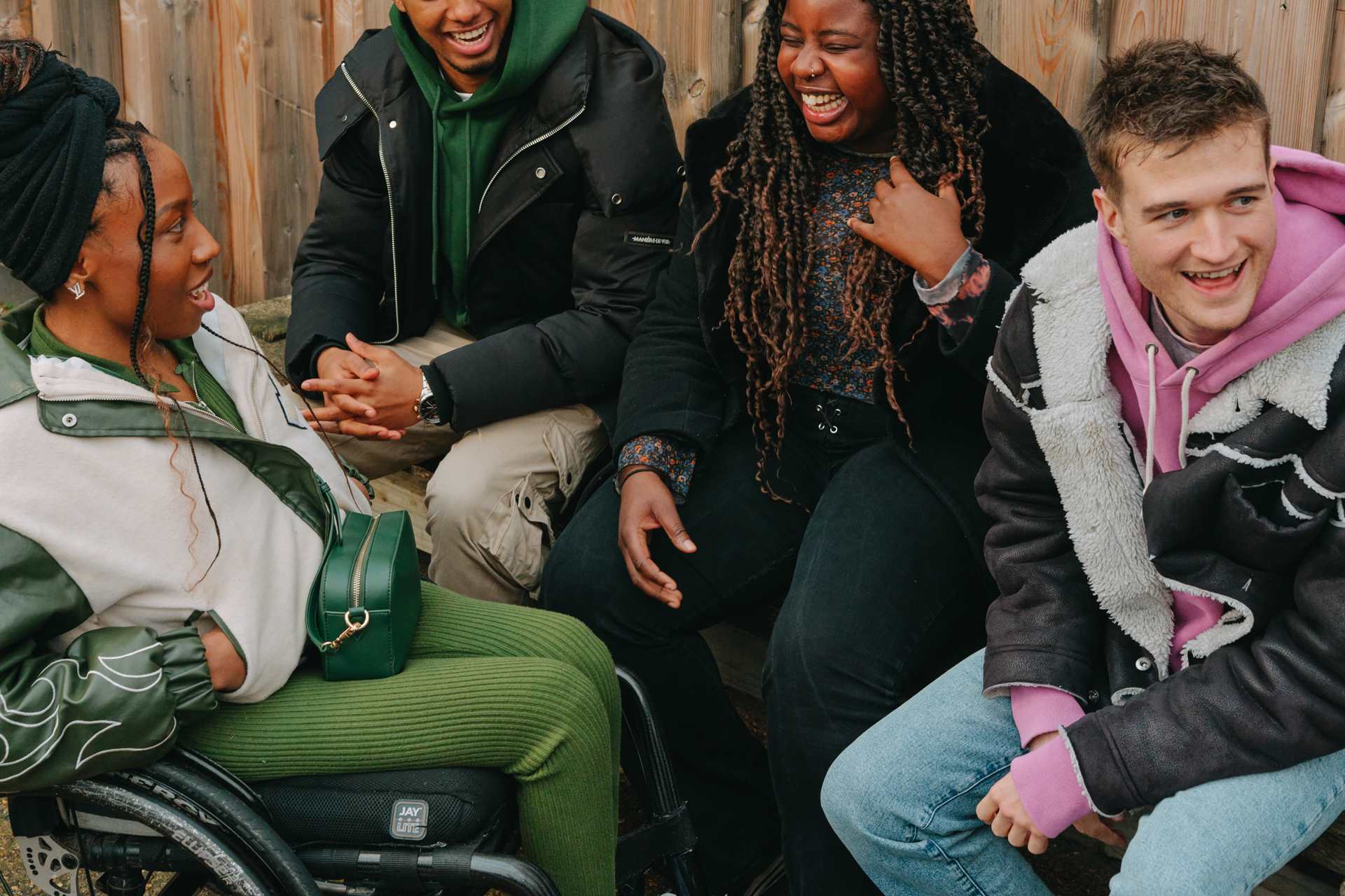 A group of young people laughing together outside on a bench. Group includes two Black young women (one in a wheelchair), one Black young man, and a white young man.