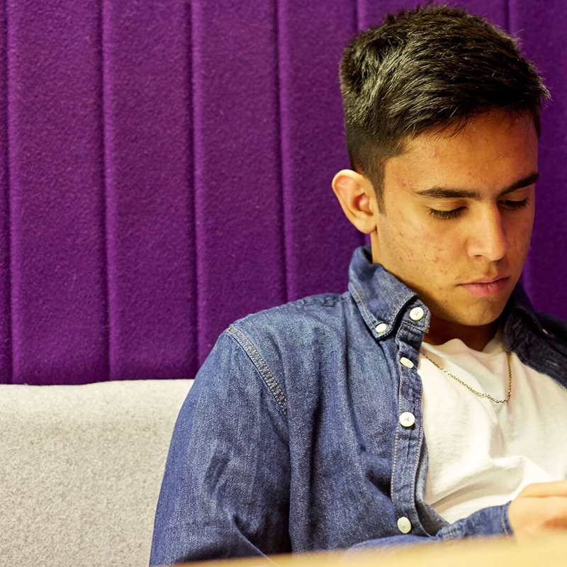 A boy in a blue denim shirt looks at his phone while sitting down against a grey sofa with a purple wall.