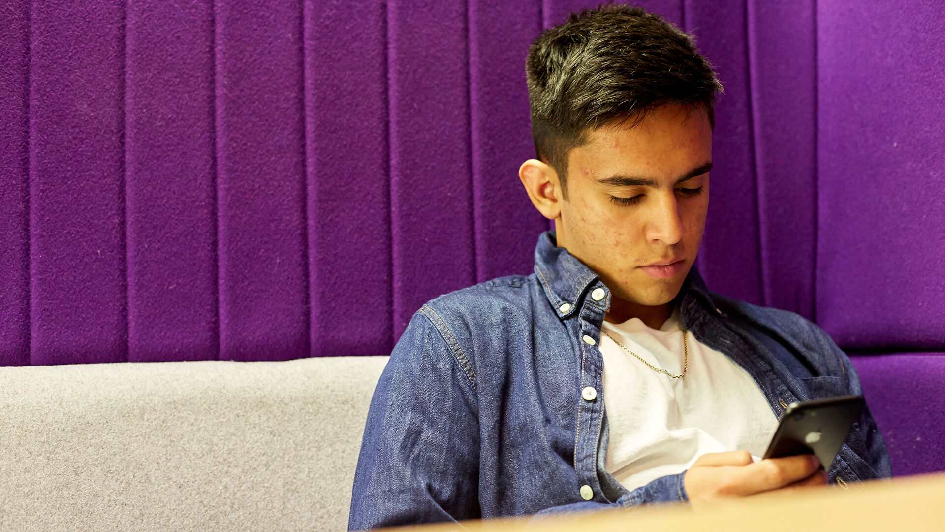 A boy in a blue denim shirt looks at his phone while sitting down against a grey sofa with a purple wall.