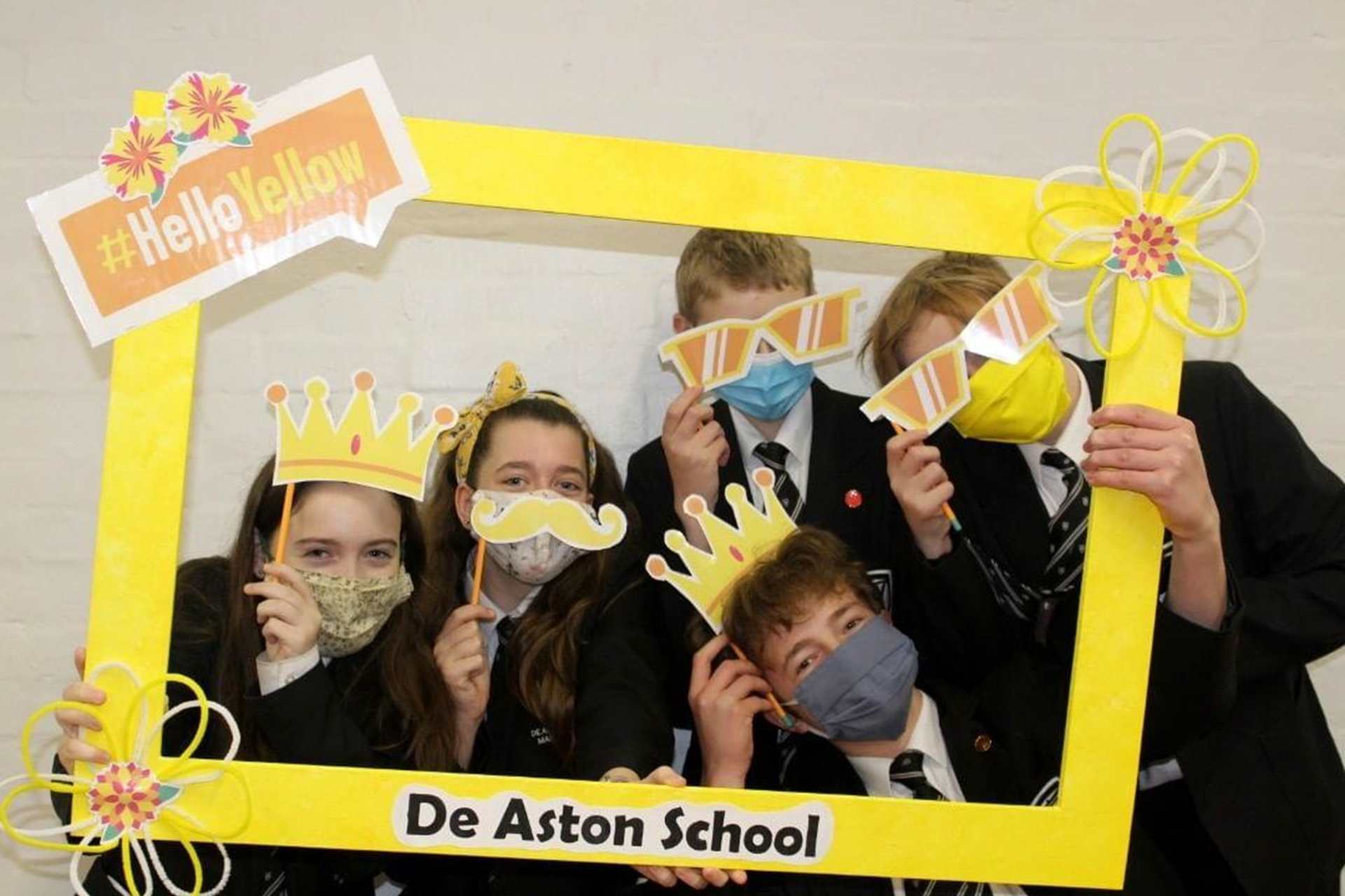 Five young people pose in a yellow photo frame with different yellow props including crowns, a yellow moustache and sunglasses. They are all wearing face masks and are facing the camera.