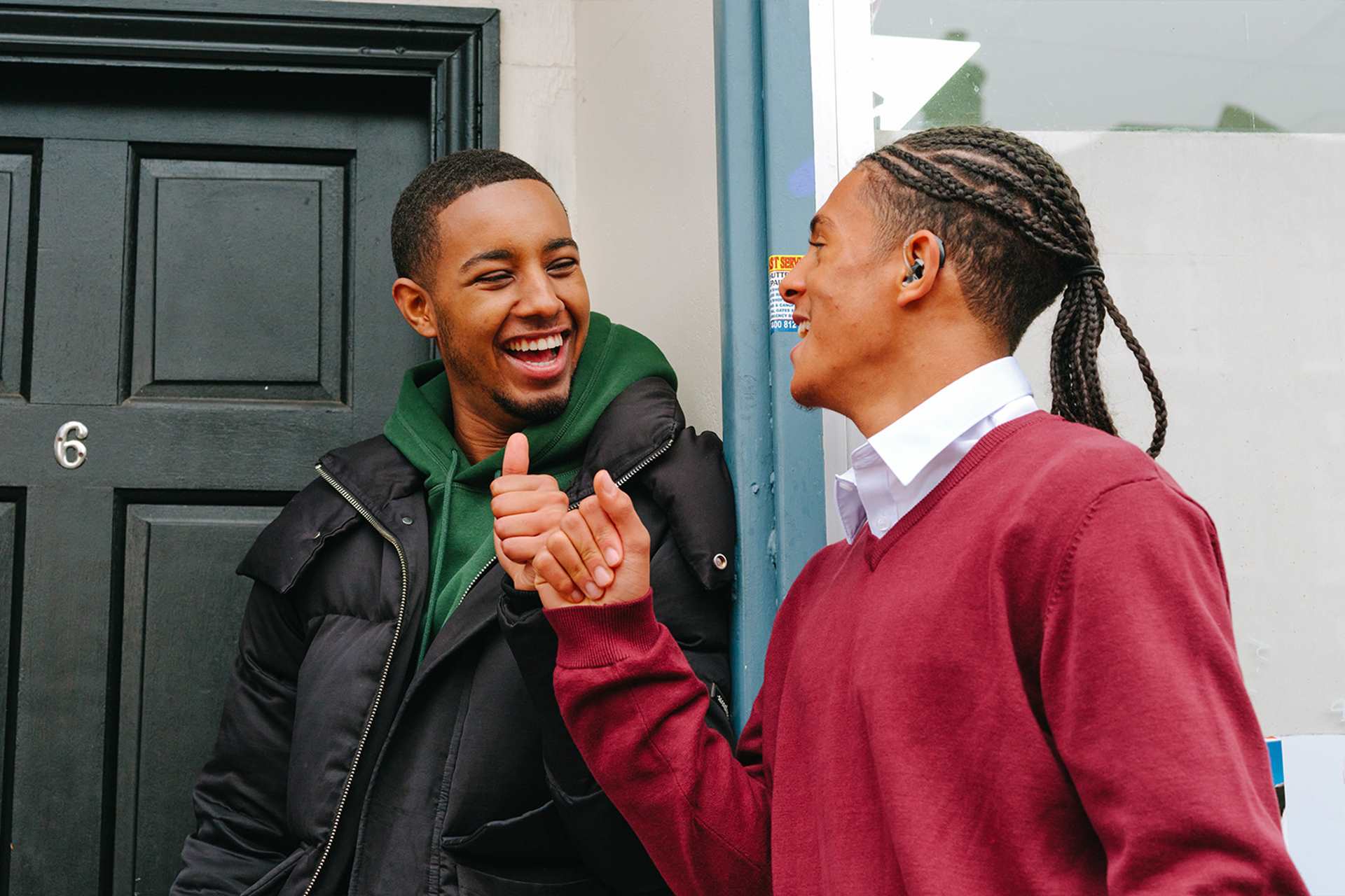 A Black teenage boy wearing a hearing aid bumping fists with a young Black man outside a front door.