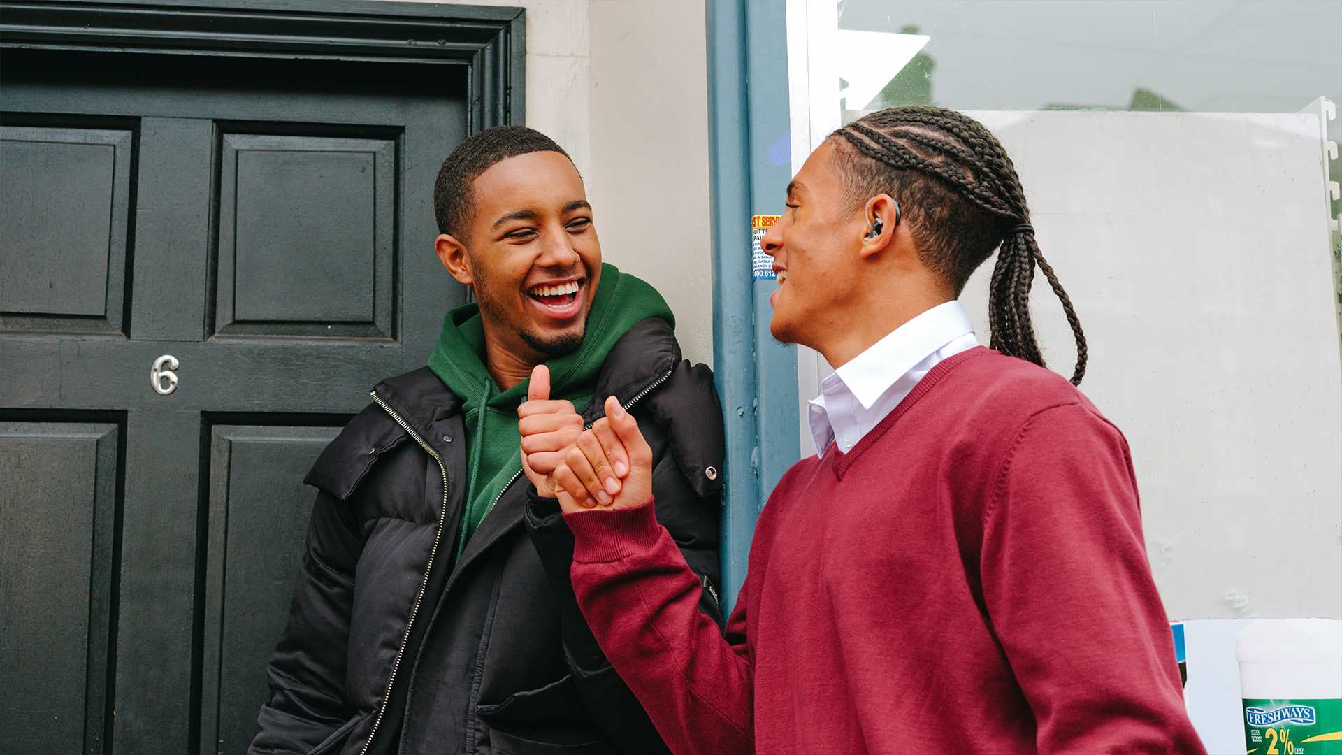 A Black teenage boy wearing a hearing aid bumping fists with a young Black man outside a front door.