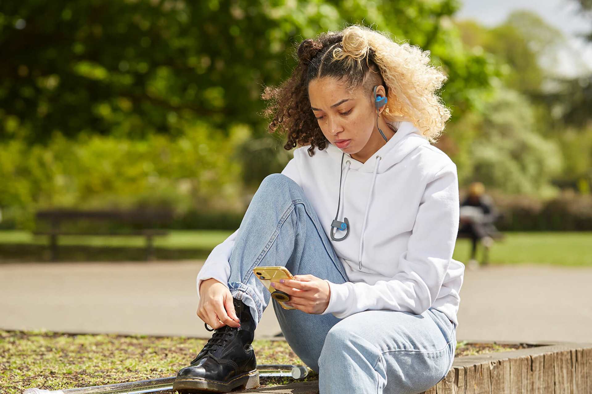 A girl sitting in the park wearing headphones. She is looking down at her phone and listening to music.