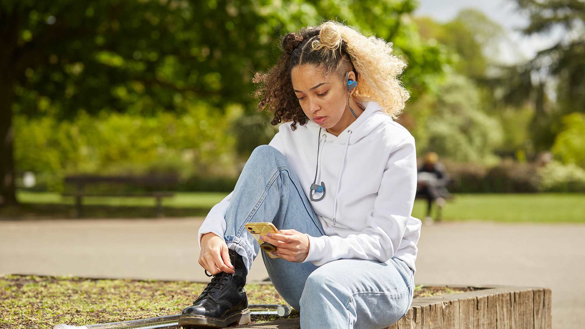 A girl sitting in the park wearing headphones. She is looking down at her phone and listening to music.