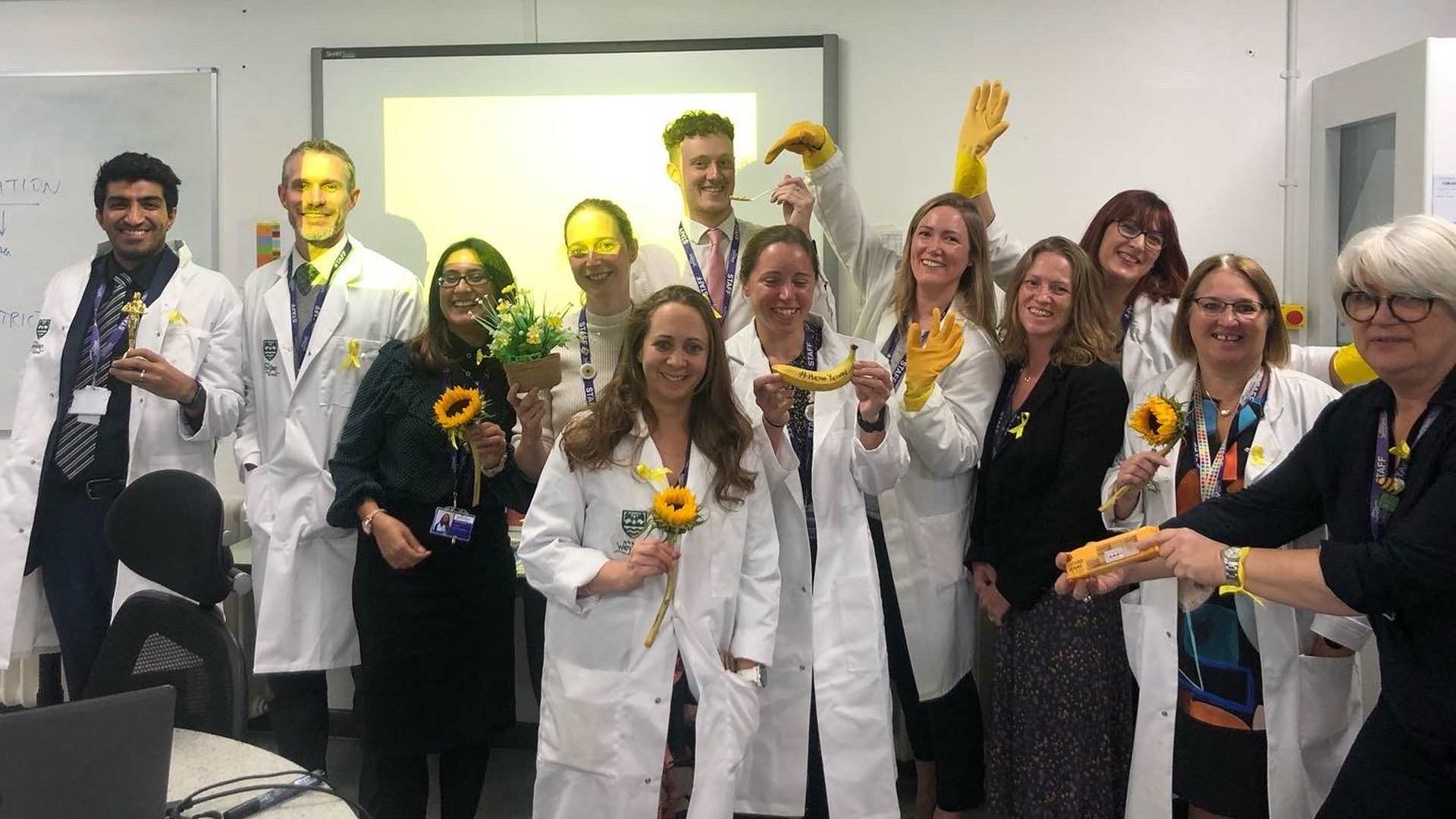 A group of teachers and staff from Weydon School, holding yellow flowers, bananas, and gloves.