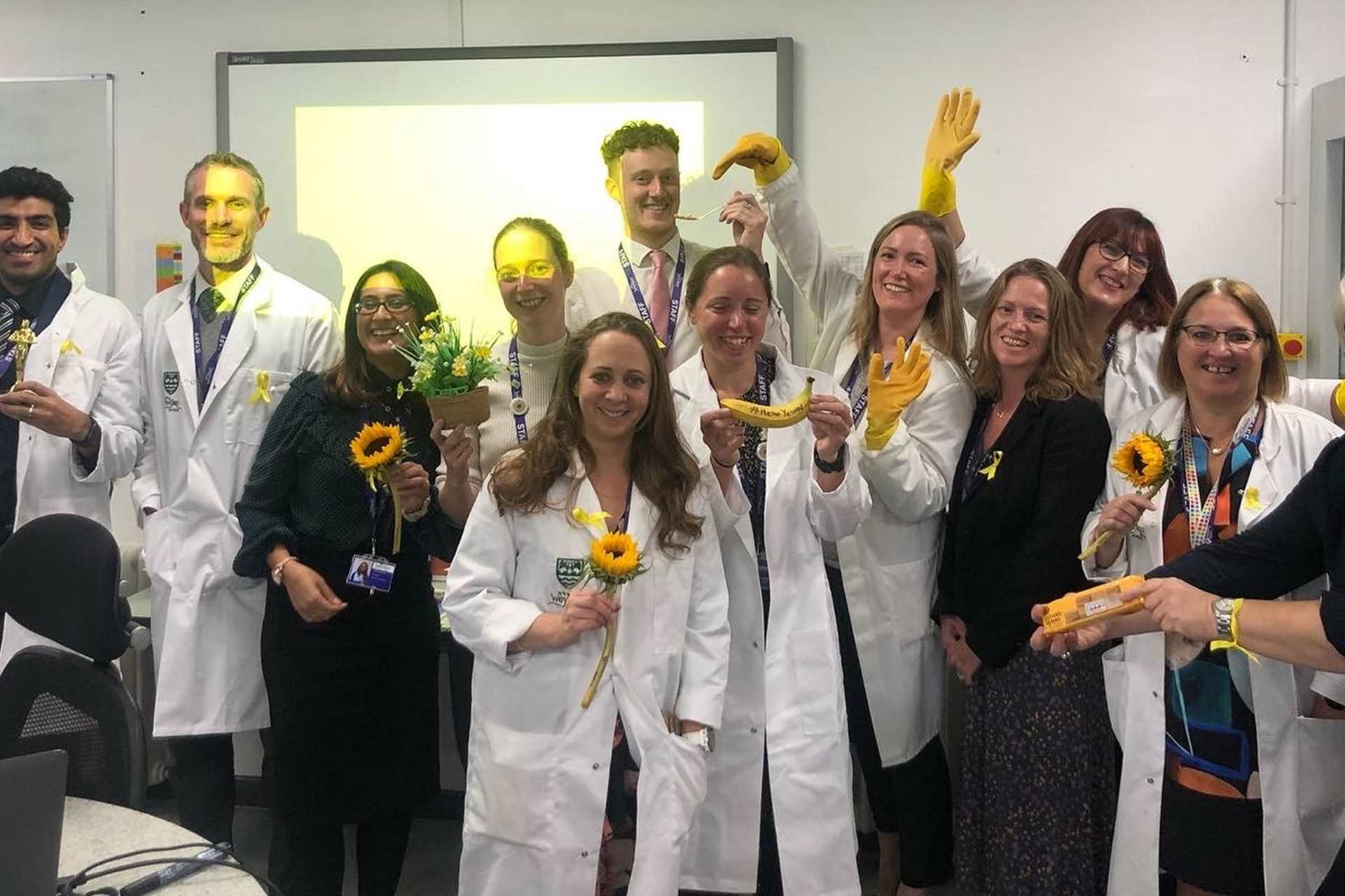 A group of teachers and staff from Weydon School, holding yellow flowers, bananas, and gloves.