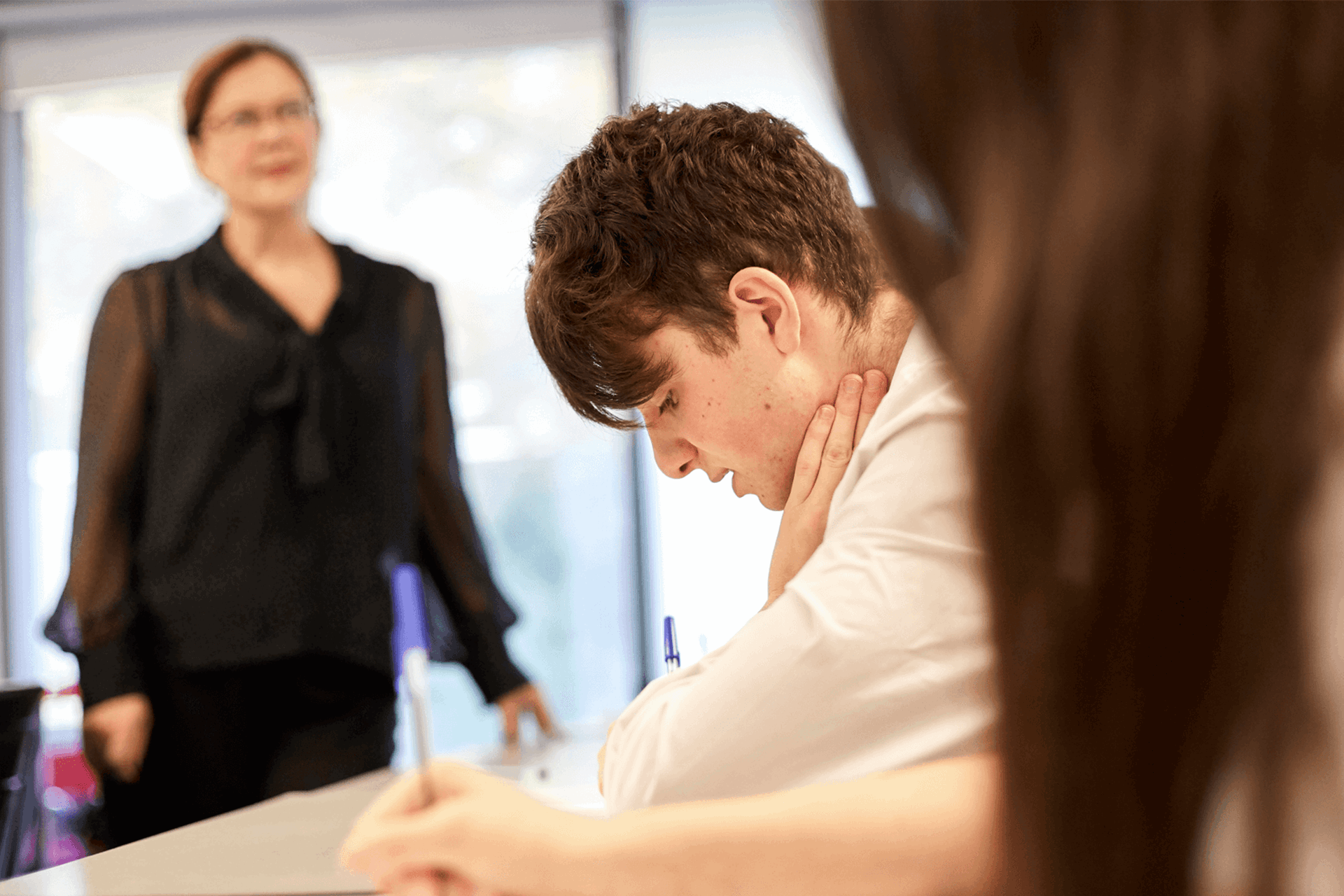 a-boy-in-a-uniform-sitting-on-his-desk-writing-on-his-desk-and-hands-on-his-neck-while-exams-is-on-going-in-class-a-teacher-is-standing-on-the-background