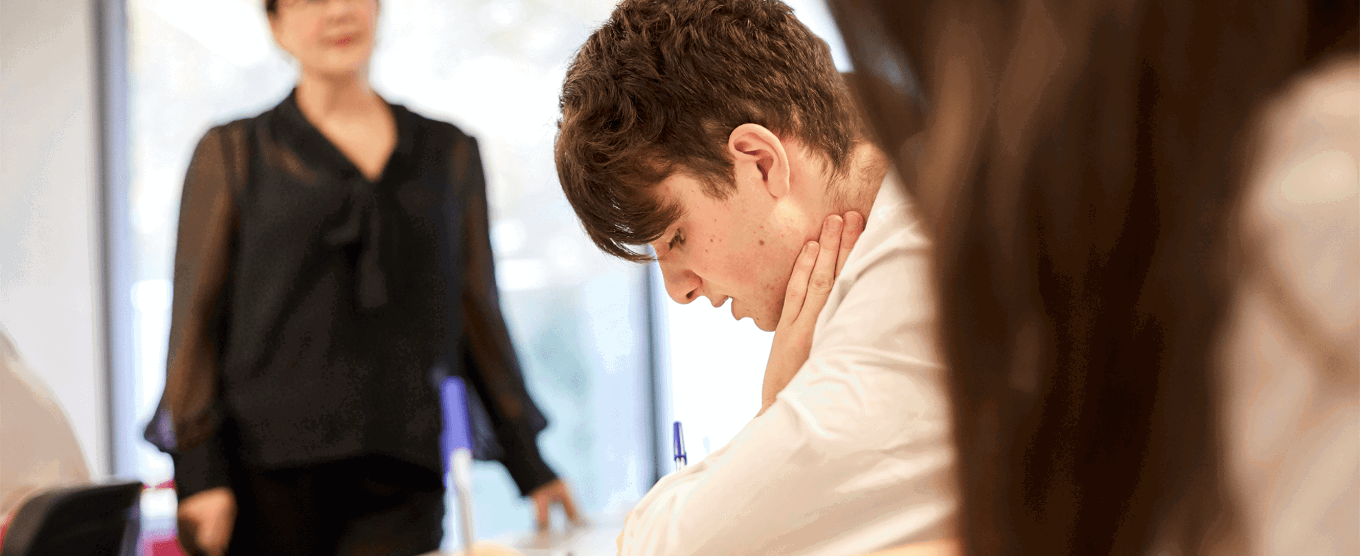 a-boy-in-a-uniform-sitting-on-his-desk-writing-on-his-desk-and-hands-on-his-neck-while-exams-is-on-going-in-class-a-teacher-is-standing-on-the-background
