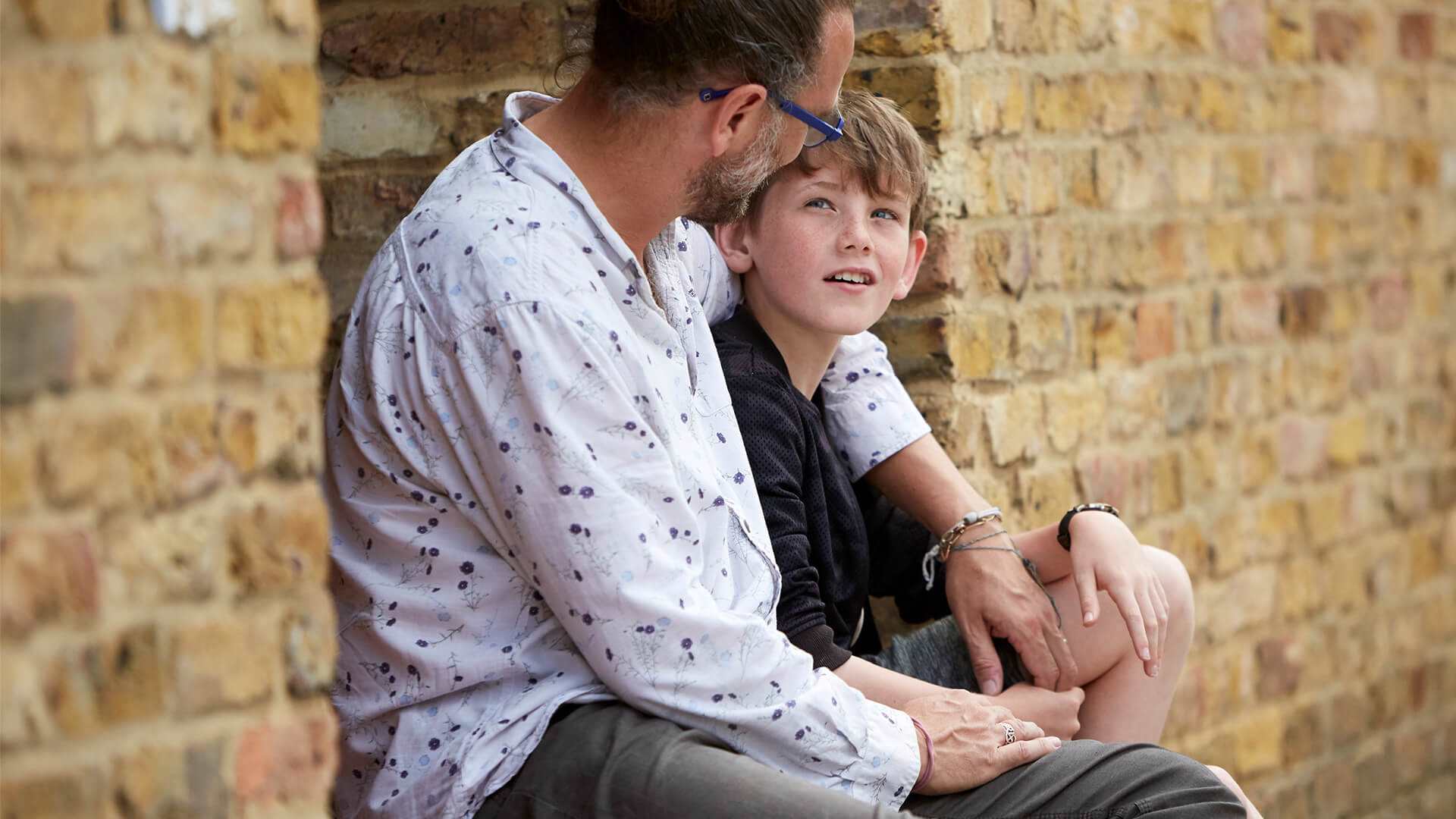 A father with his arm around his son sitting by a wall.