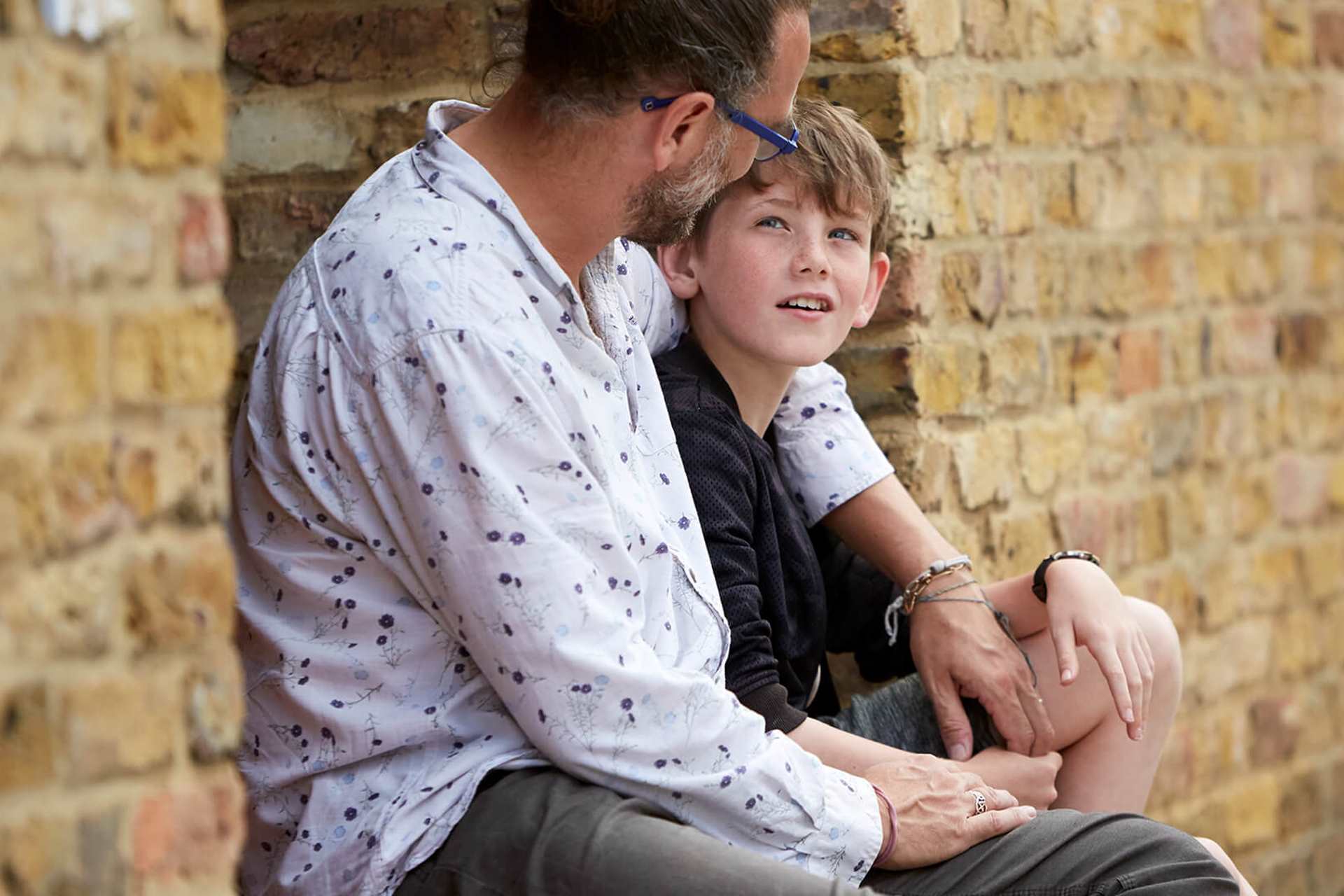 A father with his arm around his son sitting by a wall.