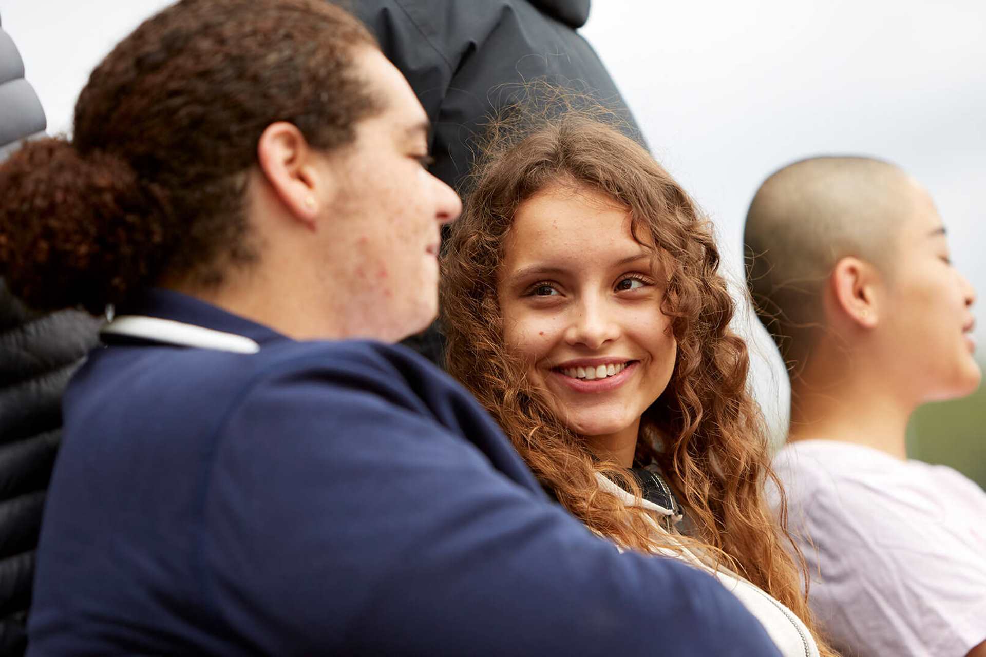 Three young people sit on a bench outside. The person on the right is chatting with the person in the middle of the bench who is looking at them and smiling. The person on the right is speaking to someone outside of the image.