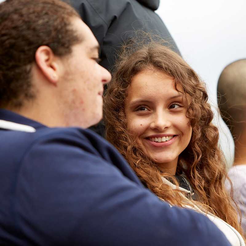 Three young people sit on a bench outside. The person on the right is chatting with the person in the middle of the bench who is looking at them and smiling. The person on the right is speaking to someone outside of the image.