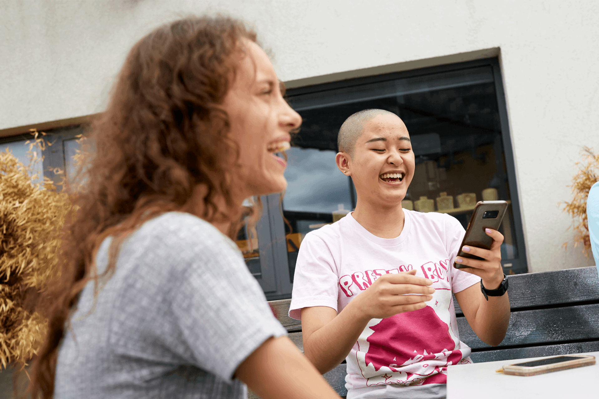 Two girls sitting together at a park bench with their friends. They are both laughing and one is looking at her phone.