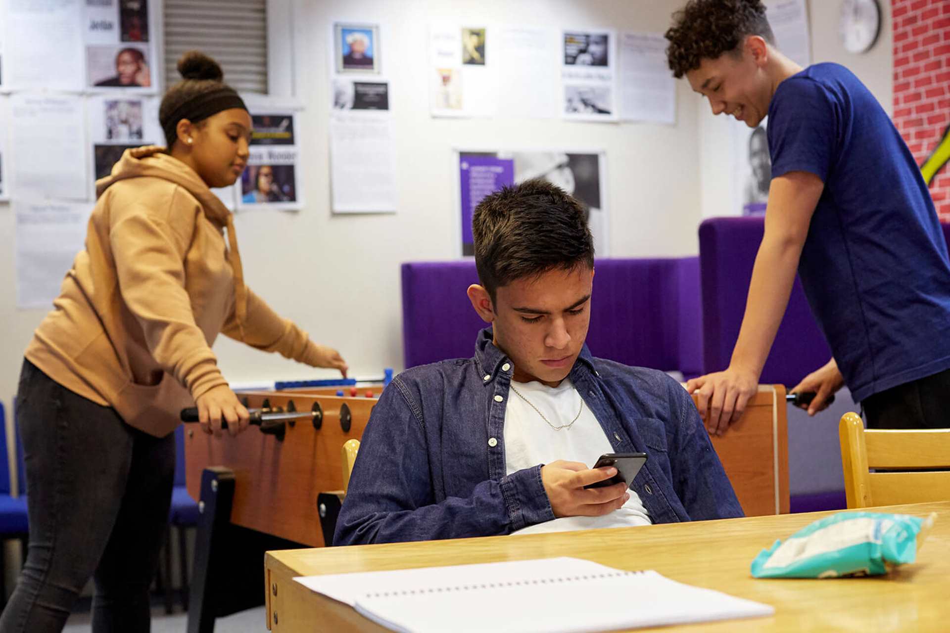 A young person is looking down at his phone at a table in a common room. There are two young people behind him playing table football.