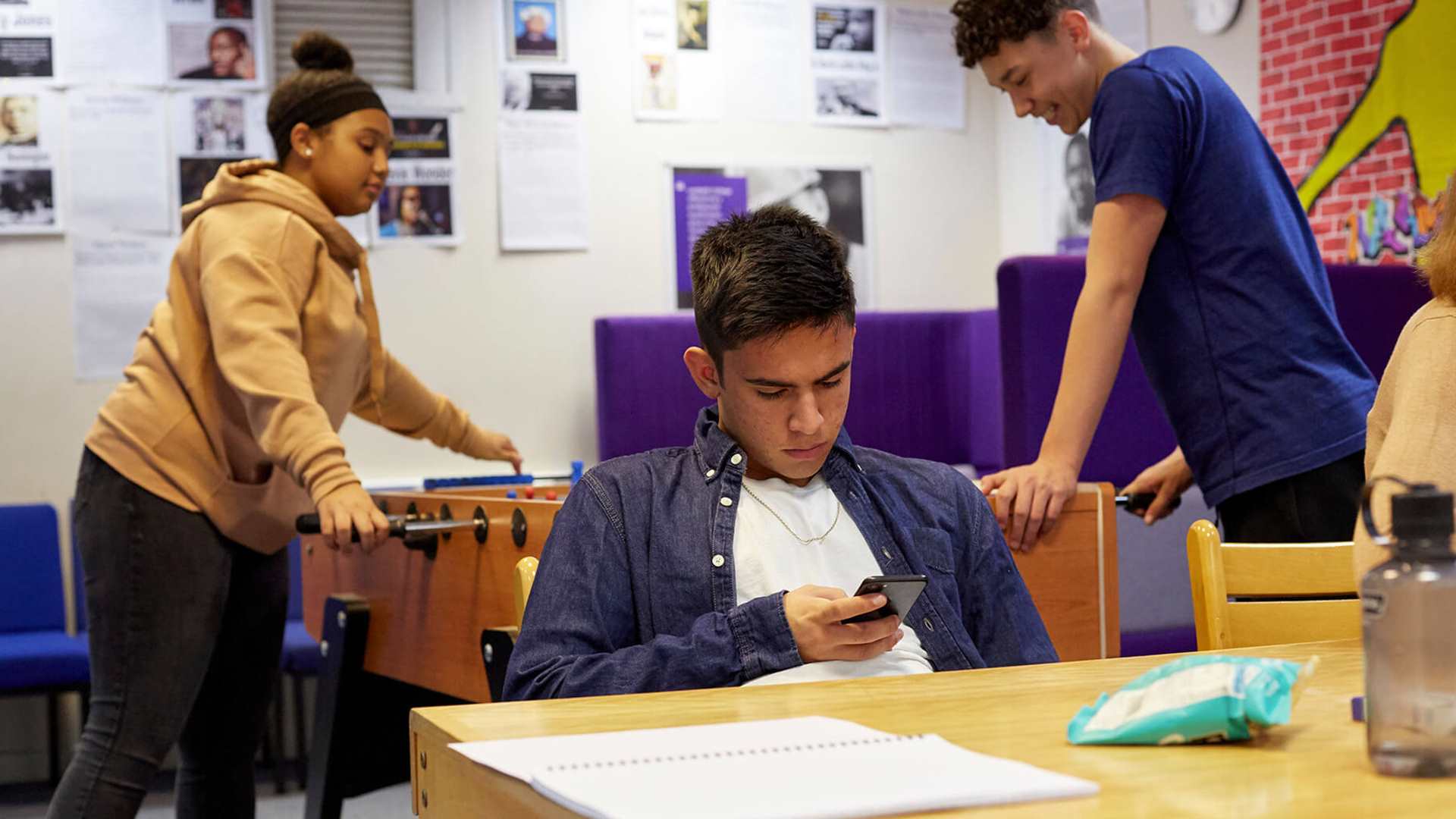 A young person is looking down at his phone at a table in a common room. There are two young people behind him playing table football.