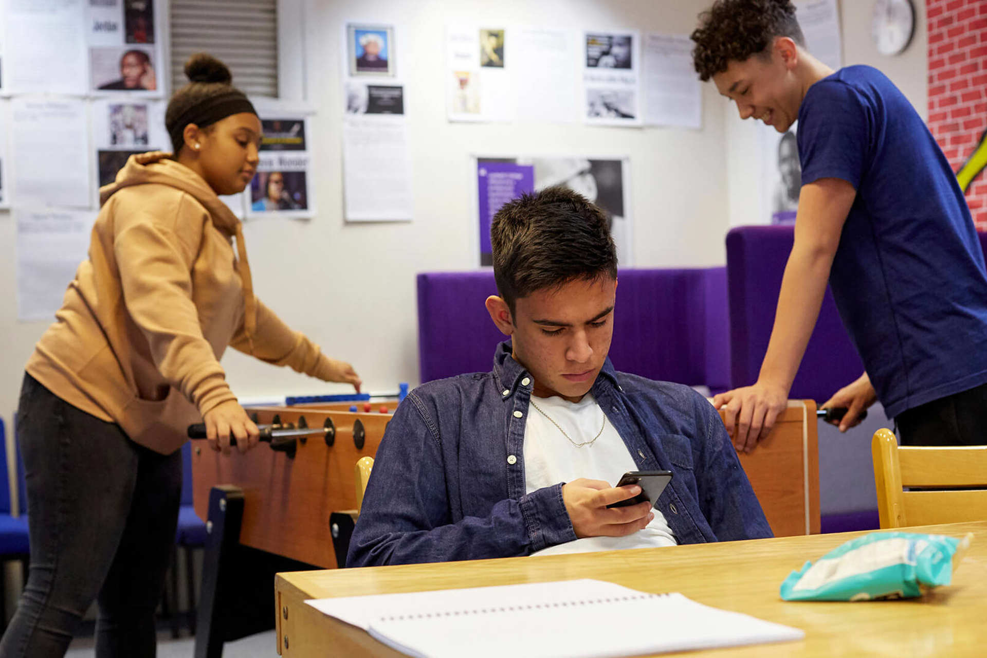 A young person is looking down at his phone at a table in a common room. There are two young people behind him playing table football.
