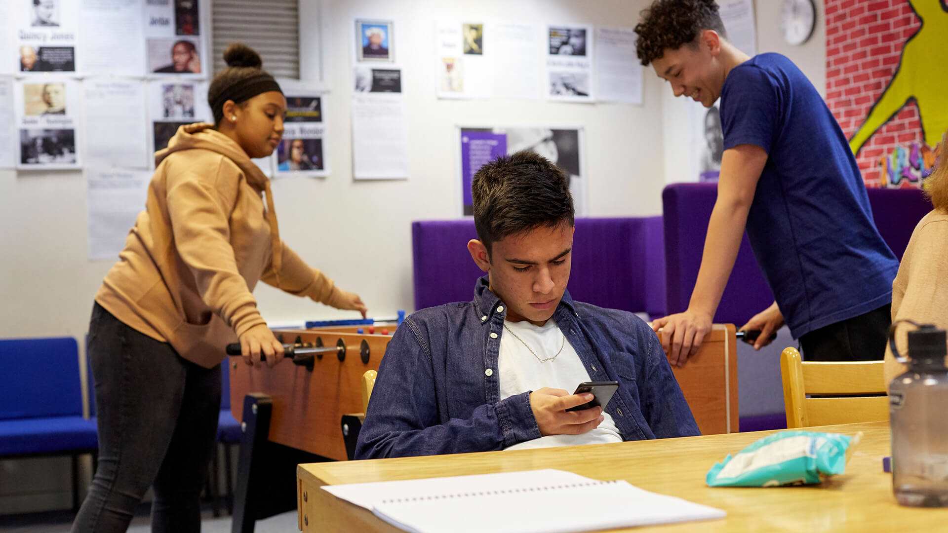 A young person is looking seriously down at his phone at a table in a common room. There are two young people behind him playing table top football, one of them is laughing.