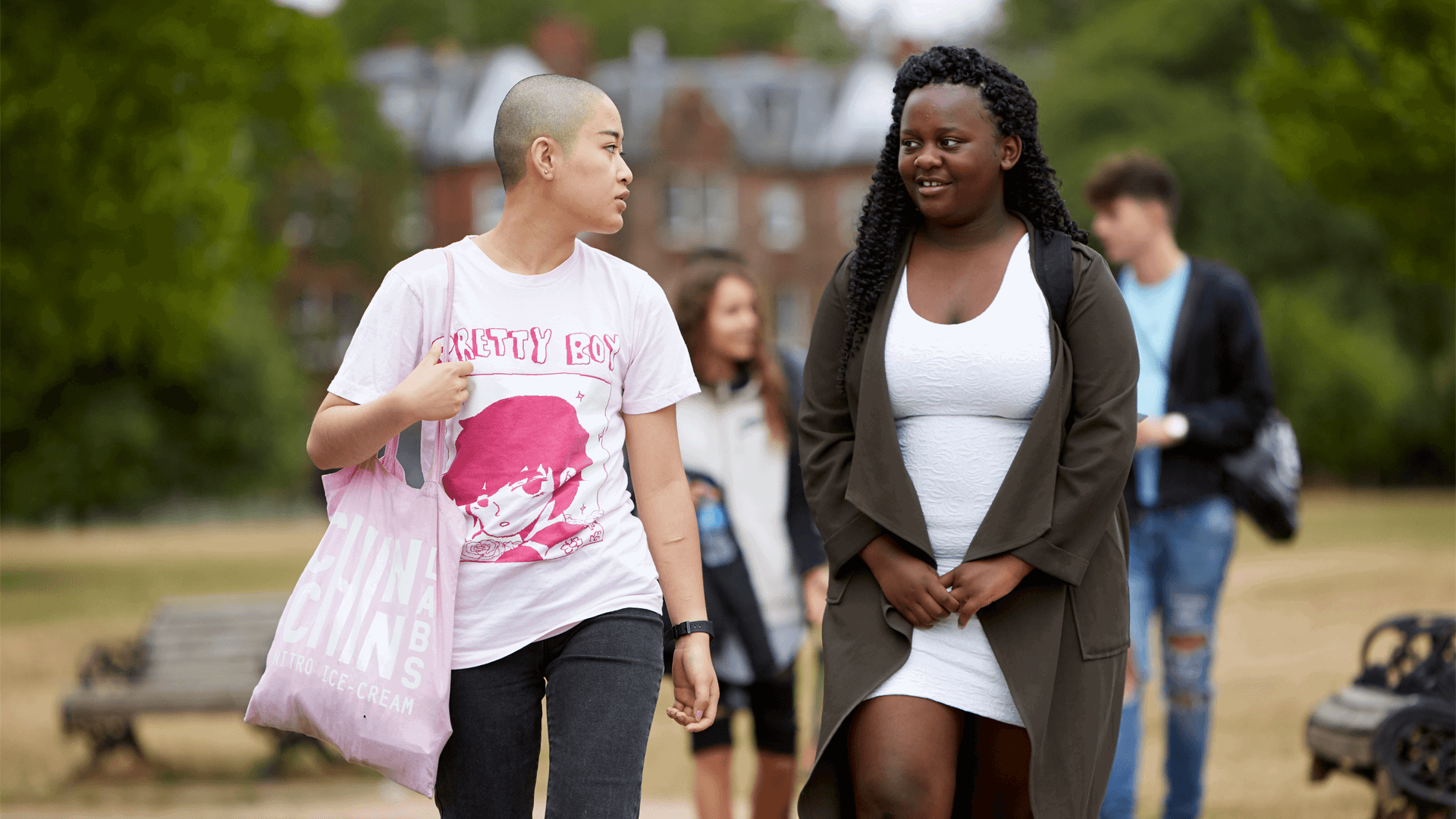 a-young-woman-with-shaved-hair-wearing-white-shirt-and-carrying-a-tote-bag-talking-to-another-young-woman-with-long-curly-hair-as-they-walk-in-a-park-with-young-people-on-their-background