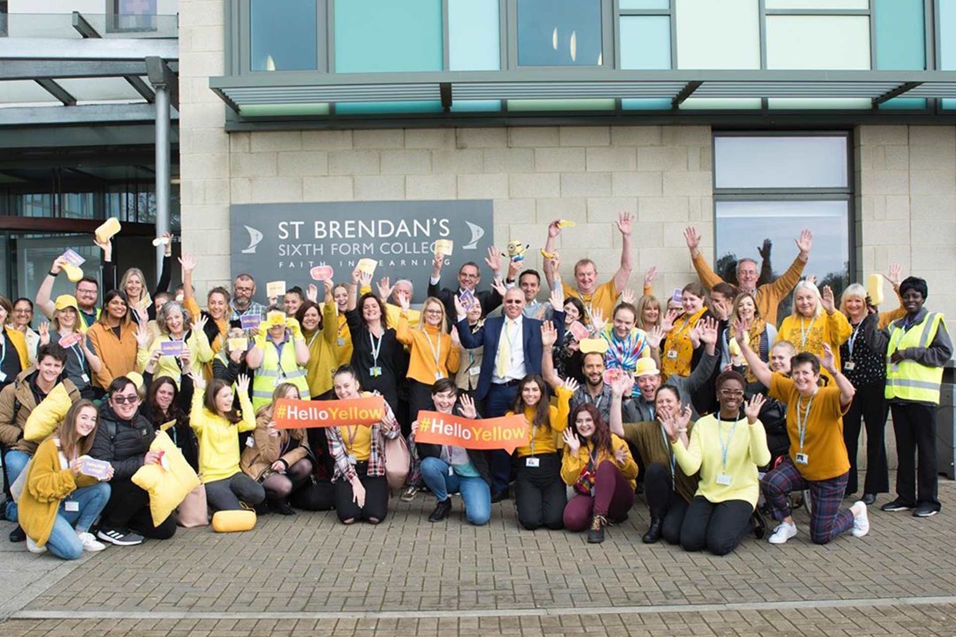 Lots of students and teachers dressed in yellow stand outside the front of their school waving and smiling at the camera. They are all wearing yellow and holding up #helloyellow signs.