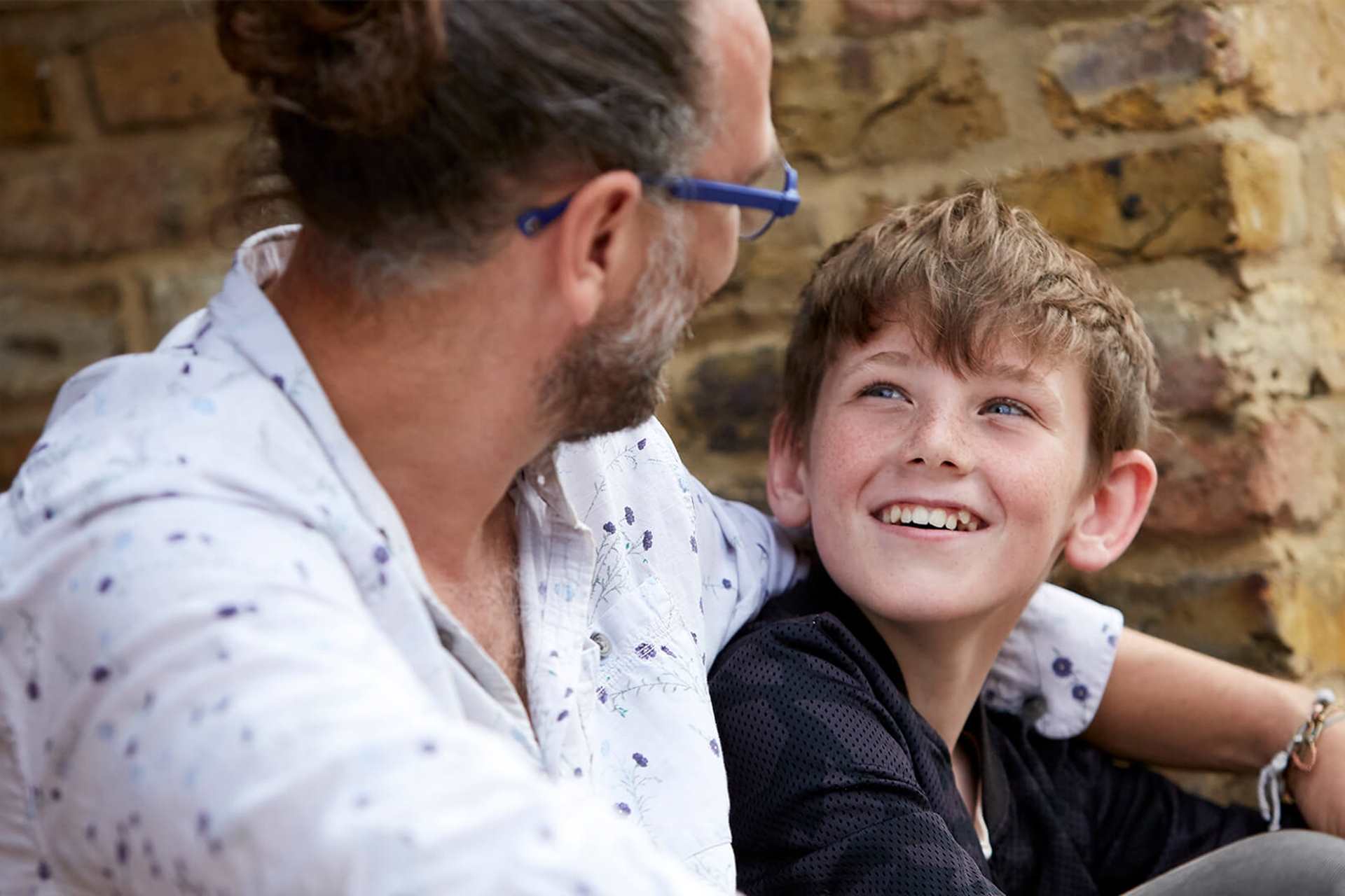 Father and son smiling and looking very happy together sitting by a wall