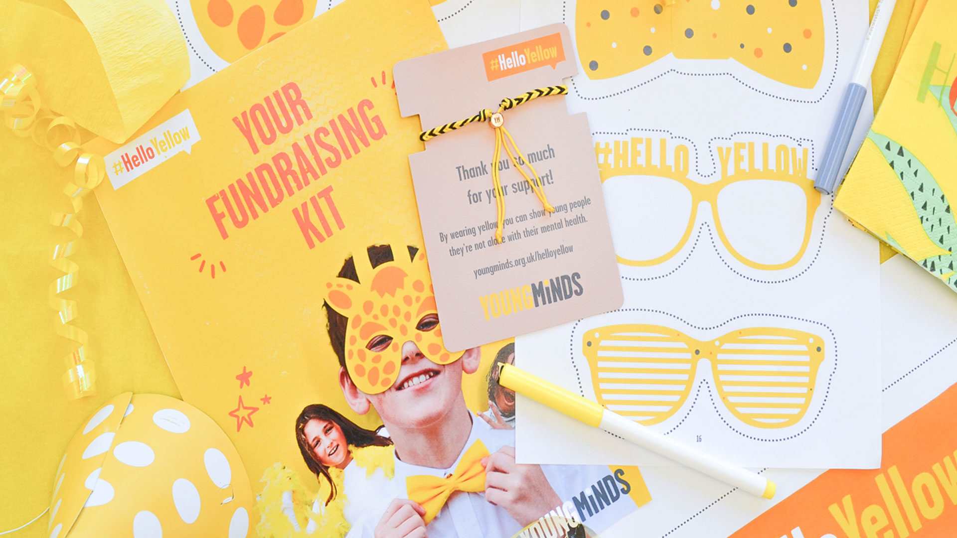 Image of Our #HelloYellow fundraising kit sits on top a yellow table. The table has pens, yellow ribbons and party hats around the kit.