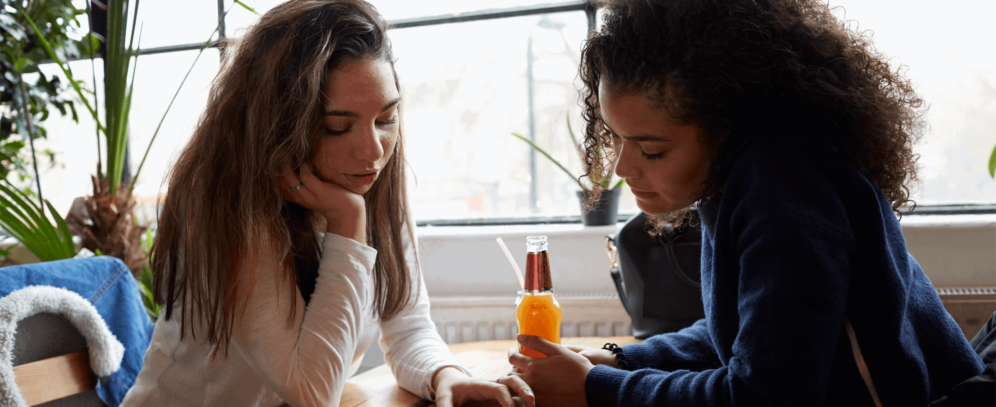 Two young women sit opposite each other at a wooden table in a restaurant. They are both looking at something on one of their smart phones, which is lying flat on the table between them,