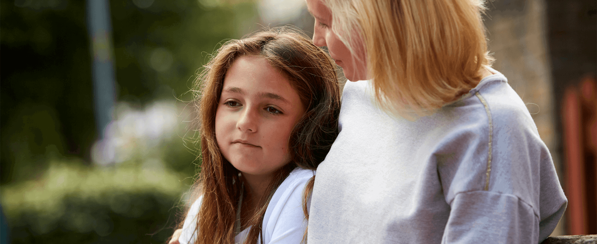 A lady comforts a young girl outside by sitting putting her arm around her