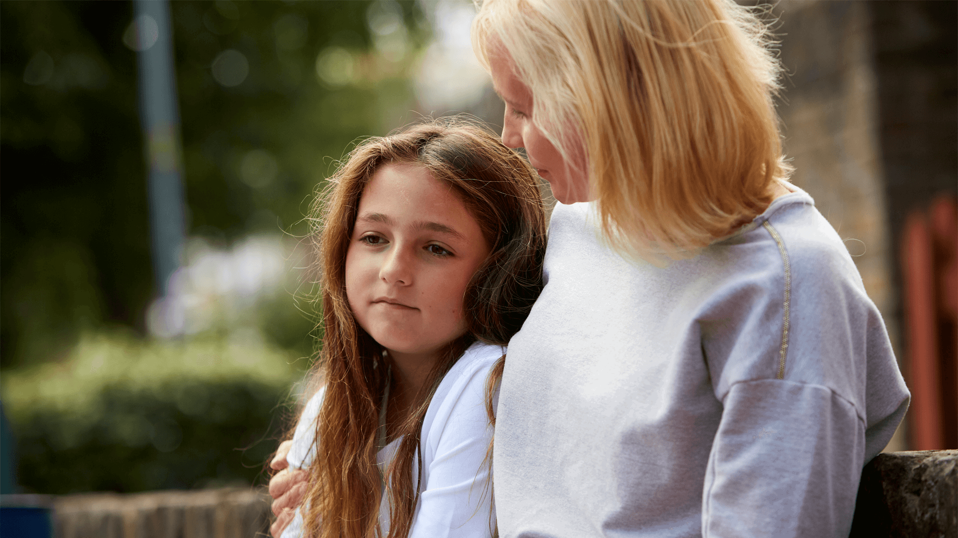 A lady comforts a young girl outside by sitting putting her arm around her