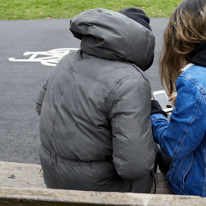 backshot of two young people wearing winter jackets with face unseen looking at their phone while sitting on a bench beside the street