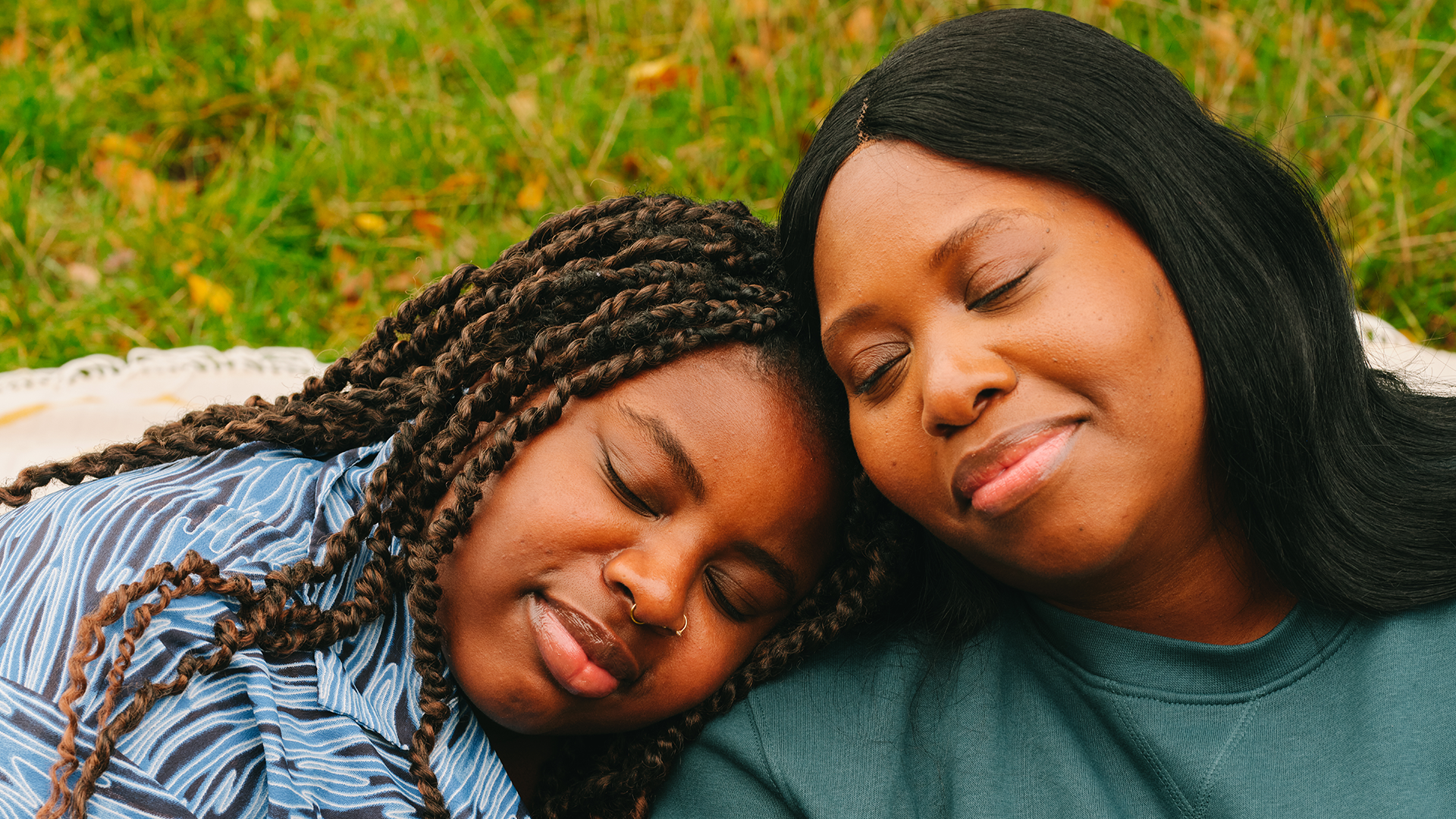 A young Black woman and an older Black woman leaning their heads on each other's shoulders and smiling. Their eyes are closed.