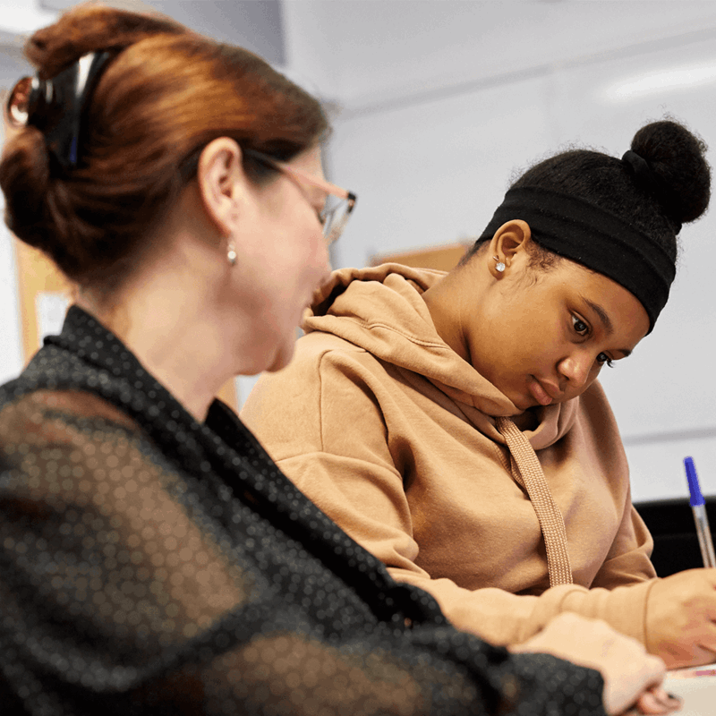 a teacher sits beside a student to help her as she answer her exam