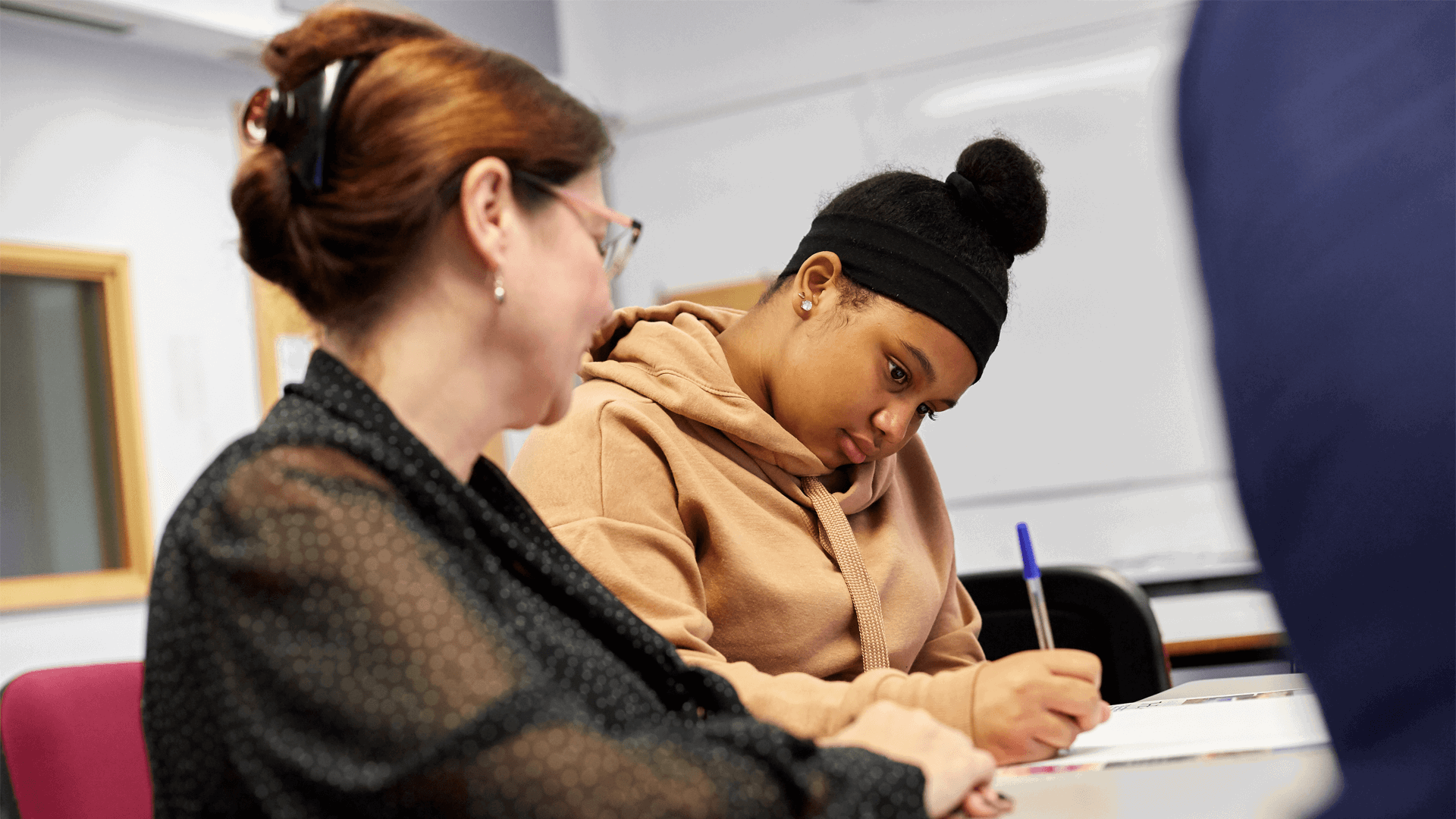 a teacher sits beside a student to help her as she answer her exam 