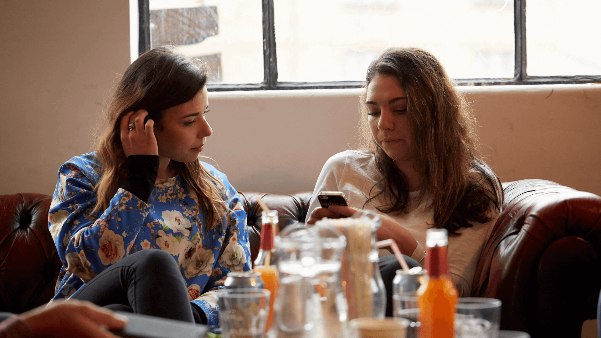medium shot of two girls sitting on a couch one is using a phone while another girl is glancing on her phone bottles and drinks are on the table as foreground